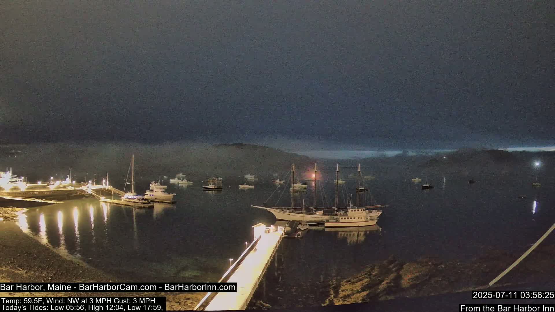 A large sailing ship and several smaller boats are moored in a calm harbor at night, under a dark, foggy sky.