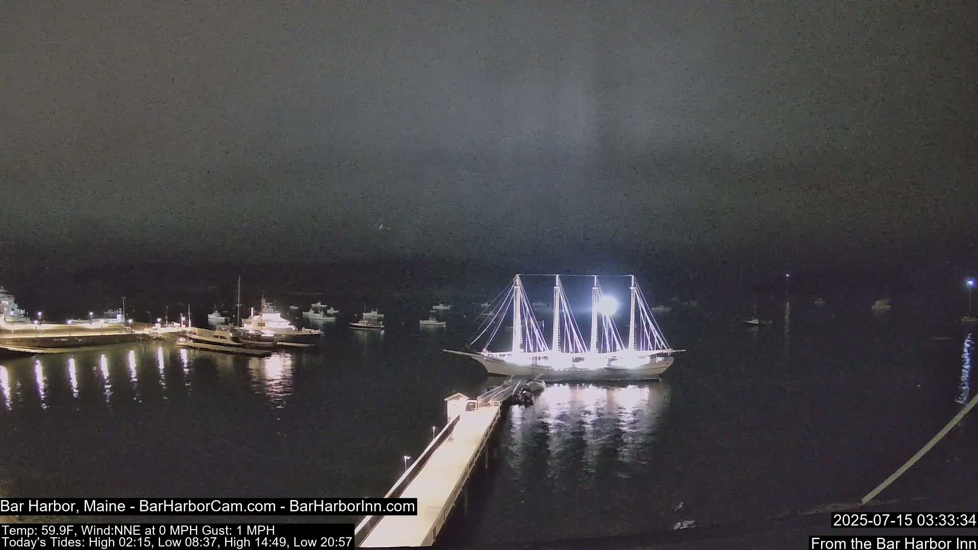 A large, brightly lit sailboat is docked near a pier at night in calm waters, with several smaller boats visible in the harbor.