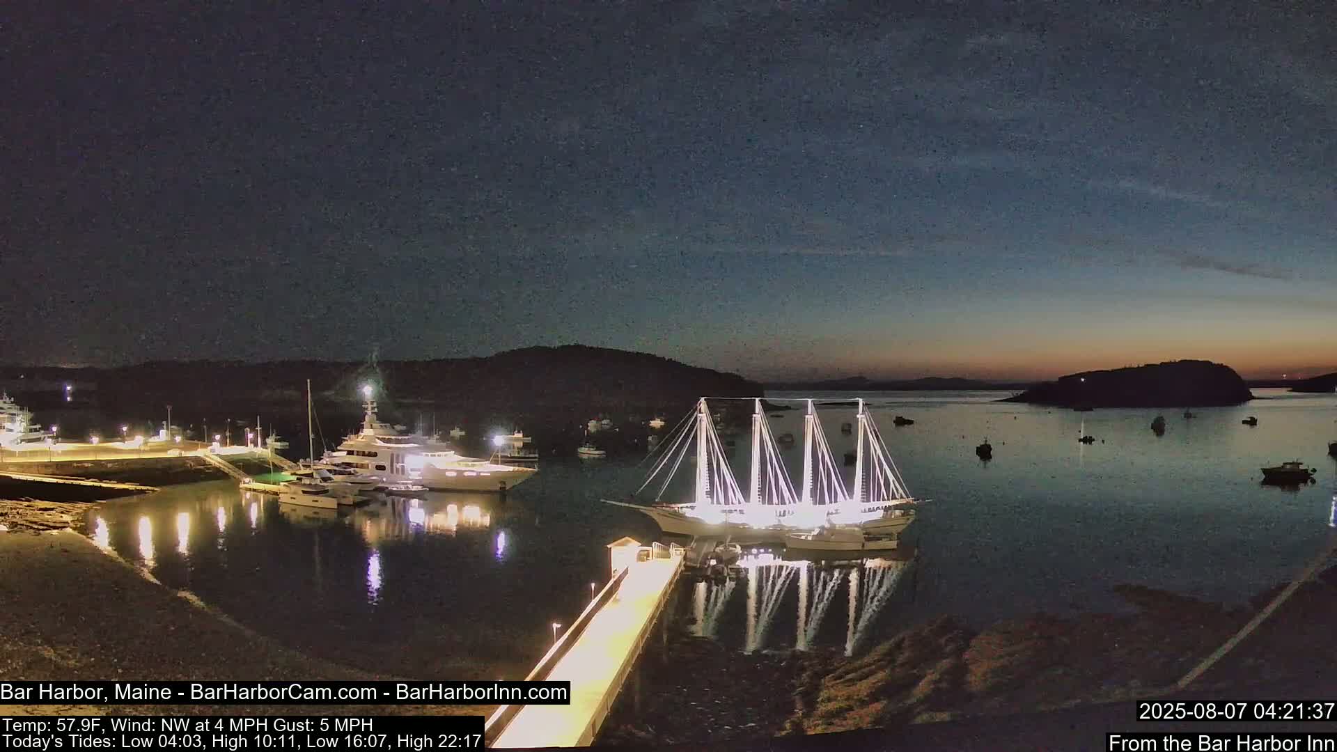 A nighttime harbor scene shows several boats, including a large illuminated sailboat docked near a pier, with calm water and a dark sky.
