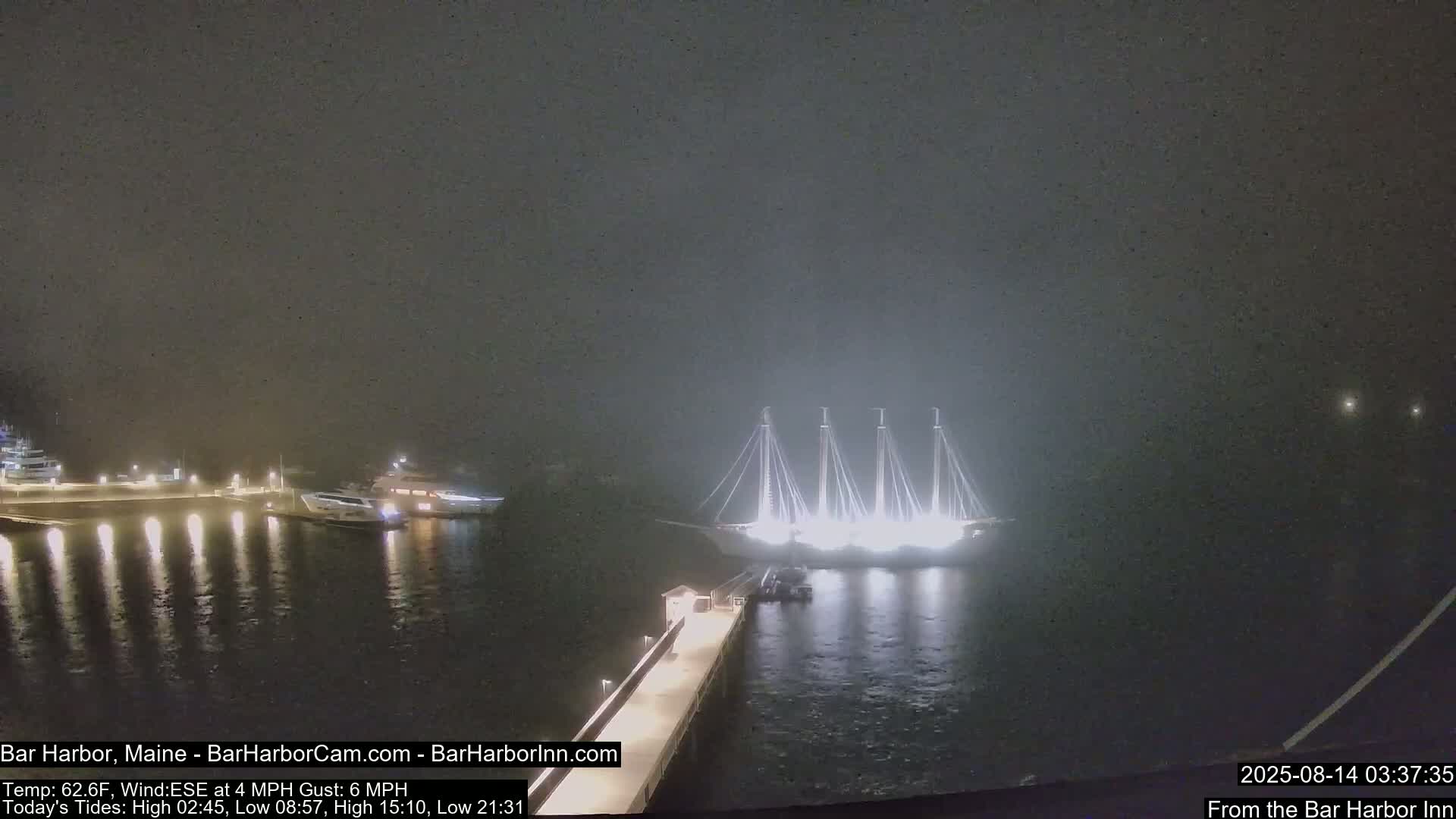 A large, brightly lit sailing ship is docked near a pier at night in foggy conditions, with smaller boats visible in the background.