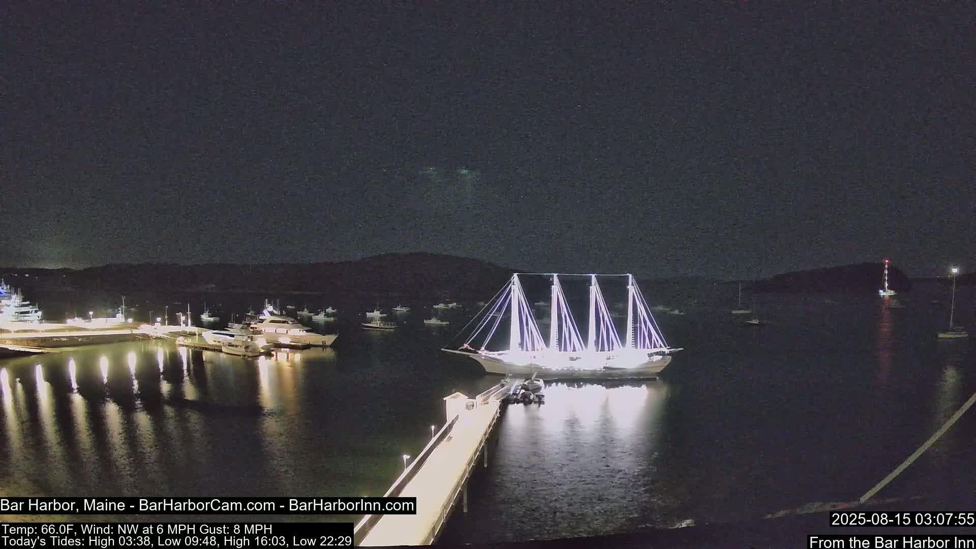 A large, brightly lit sailing ship is docked at a pier at night, with other boats in a harbor under a clear, dark sky.
