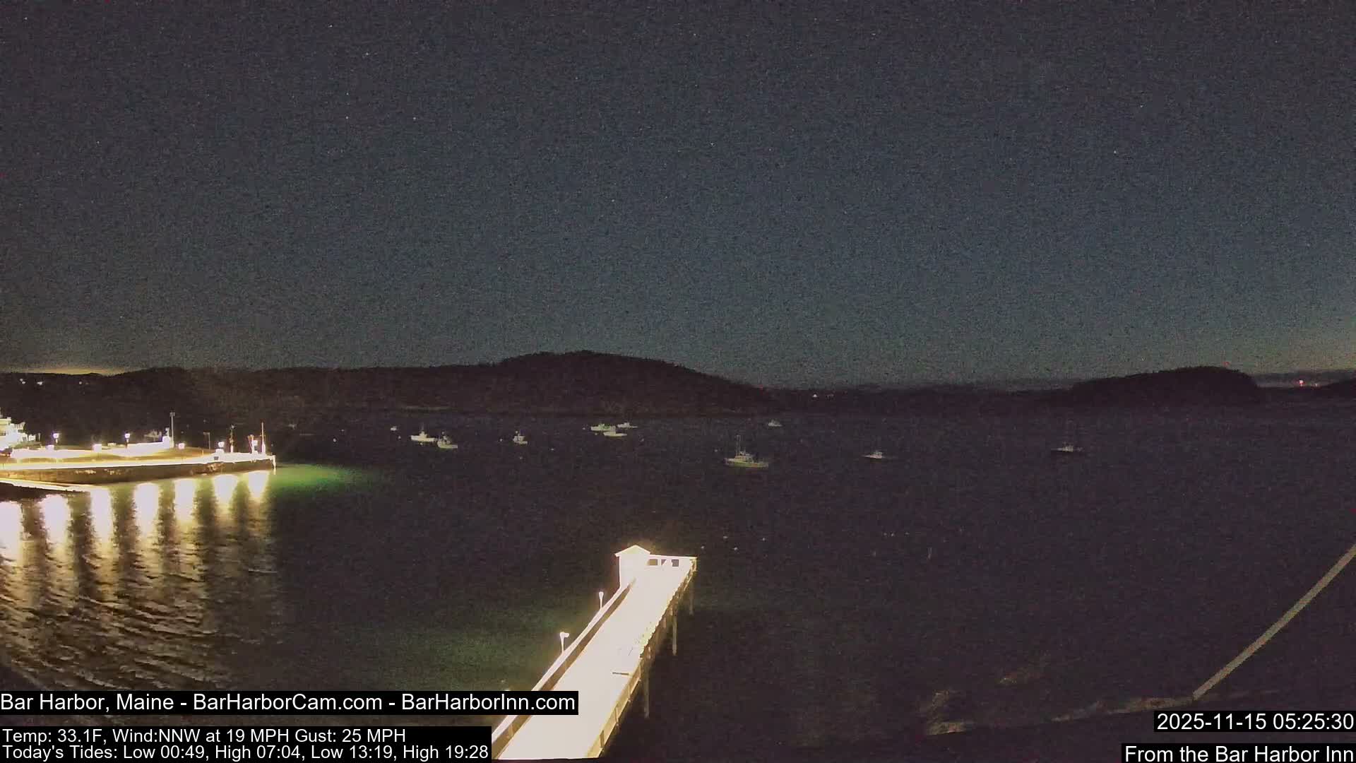 A clear, starry night illuminates a dark harbor featuring numerous anchored boats, two brightly lit piers with reflections in the calm water, and distant hills silhouetted against the horizon.