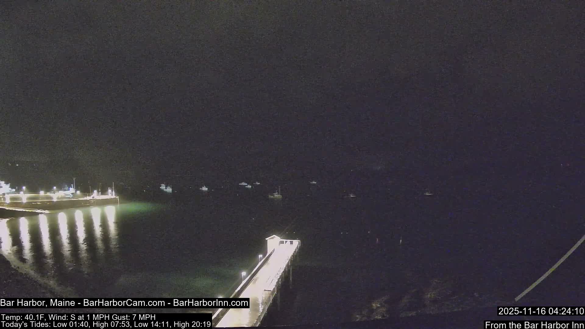 A dark, clear night reveals a brightly lit pier extending into calm water, reflecting its lights, with several boats anchored offshore under a vast, dark sky.