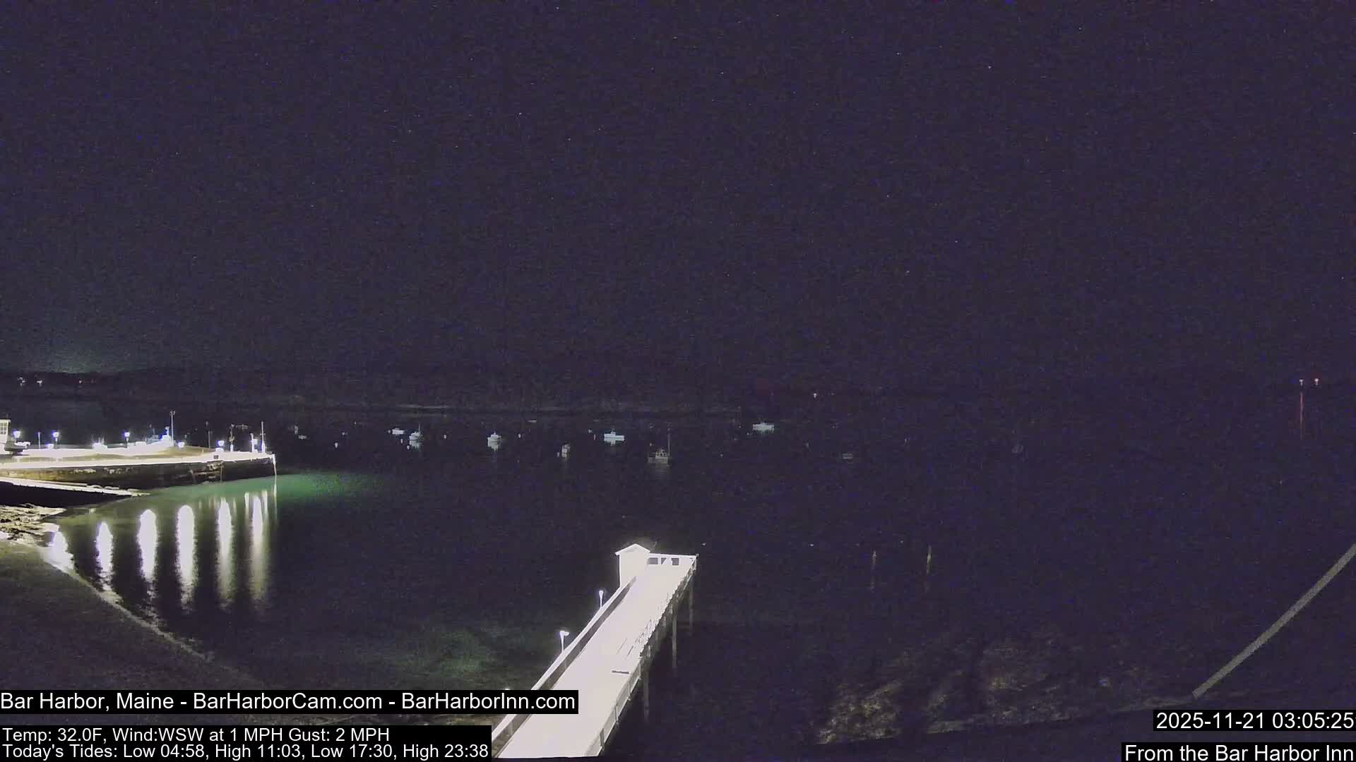 A clear, starry night overlooks a peaceful harbor featuring two illuminated piers and several boats anchored in the calm water.