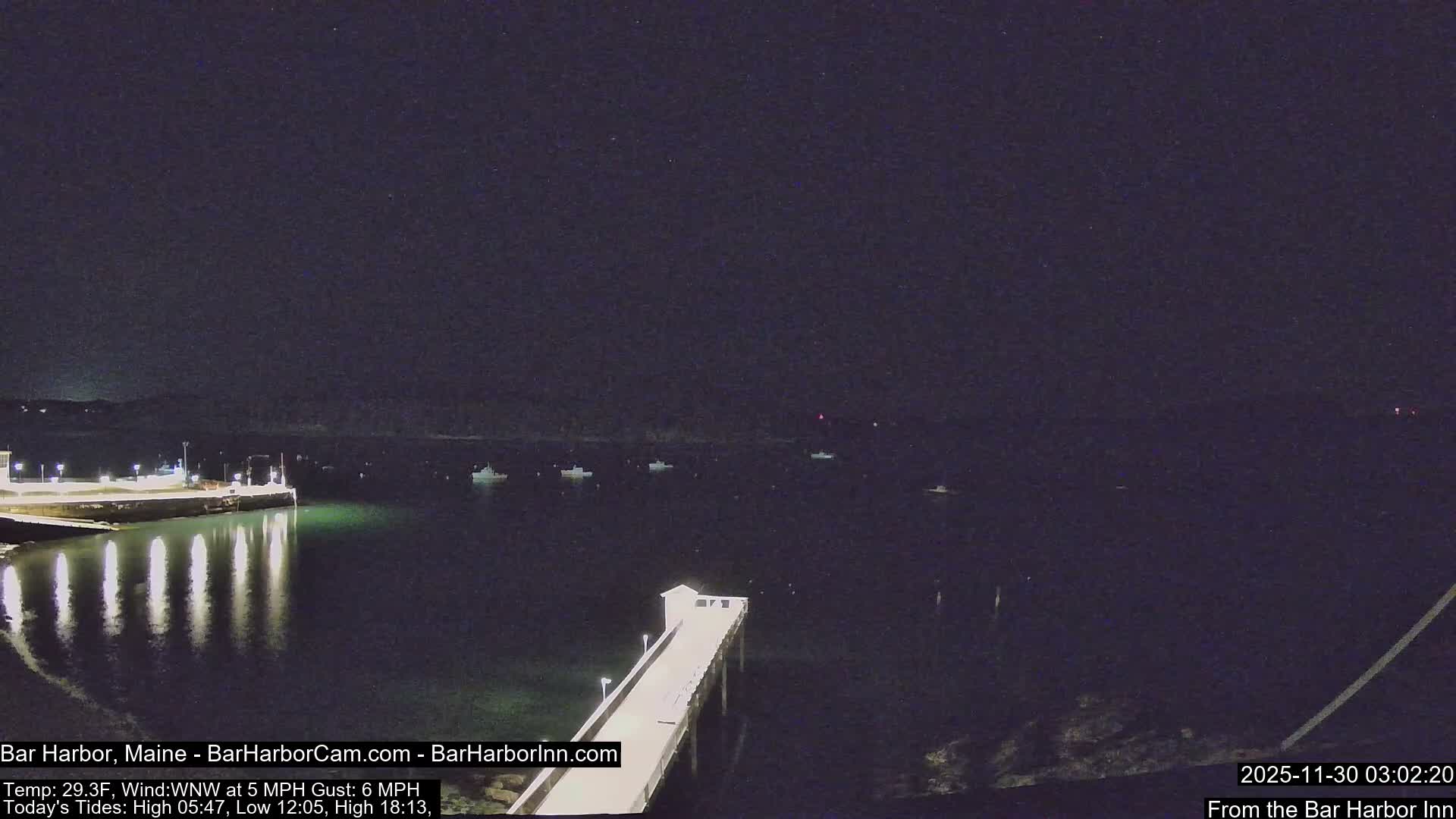 A nighttime view reveals a lit pier extending into calm, dark harbor waters reflecting the lights, with several boats anchored and distant shorelines under a clear, cold sky.