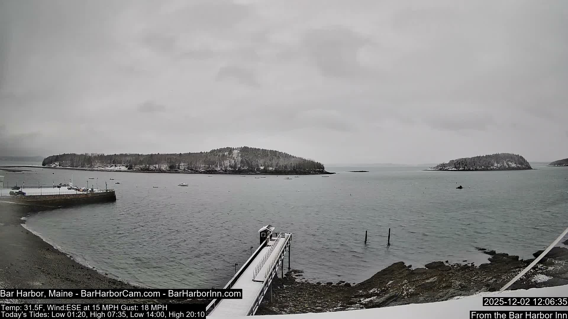 On a grey, overcast winter day, a snowy shoreline and pier lead into a calm bay with several boats, backed by snow-dusted, forested islands.