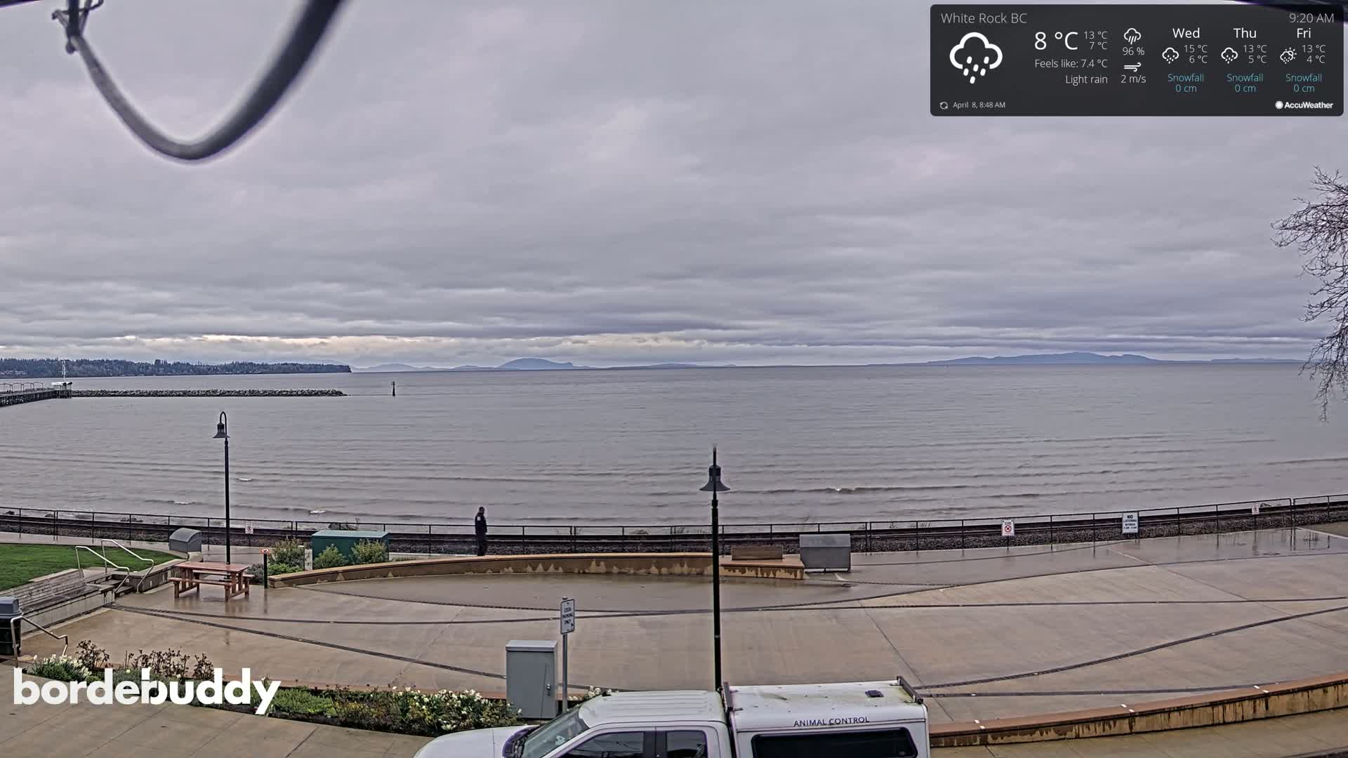 A mostly cloudy, rainy day overlooks a calm ocean with a shoreline featuring a paved area, park benches, and a rail line.