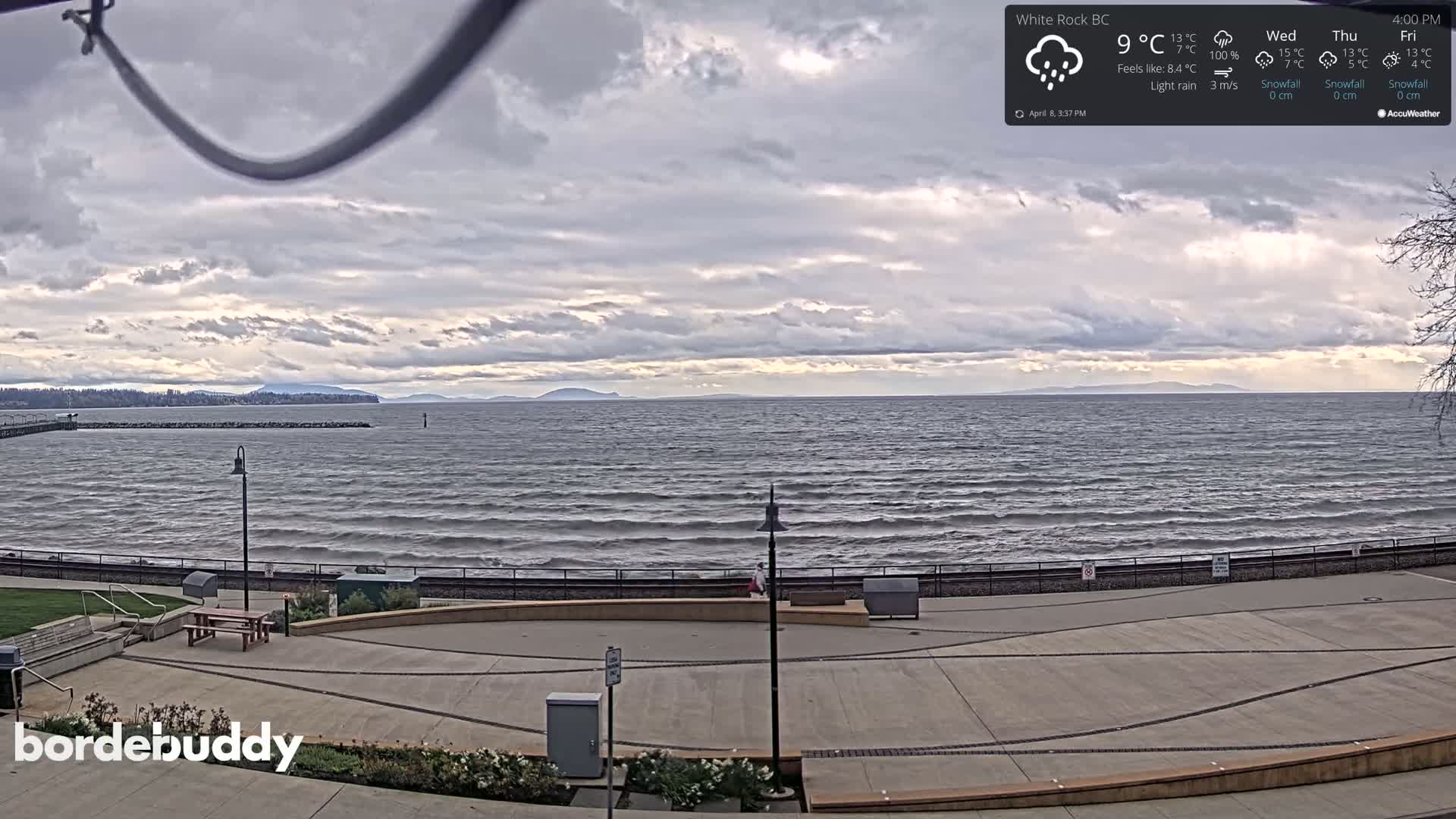A mostly cloudy day with light rain shows a view of a calm ocean with a distant shoreline and a paved waterfront area with benches and landscaping.