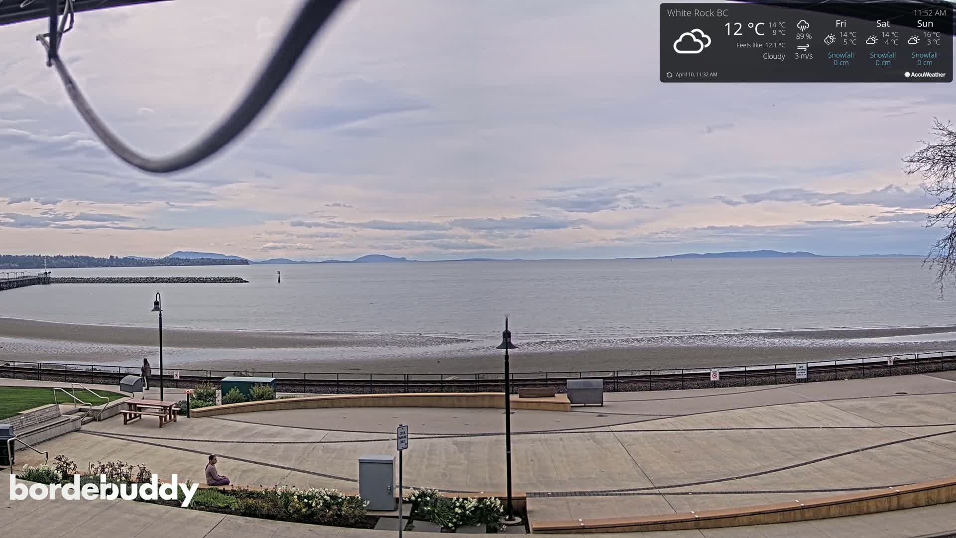 A cloudy day overlooks a calm ocean beach with a paved plaza in the foreground and a person sitting on the ground.