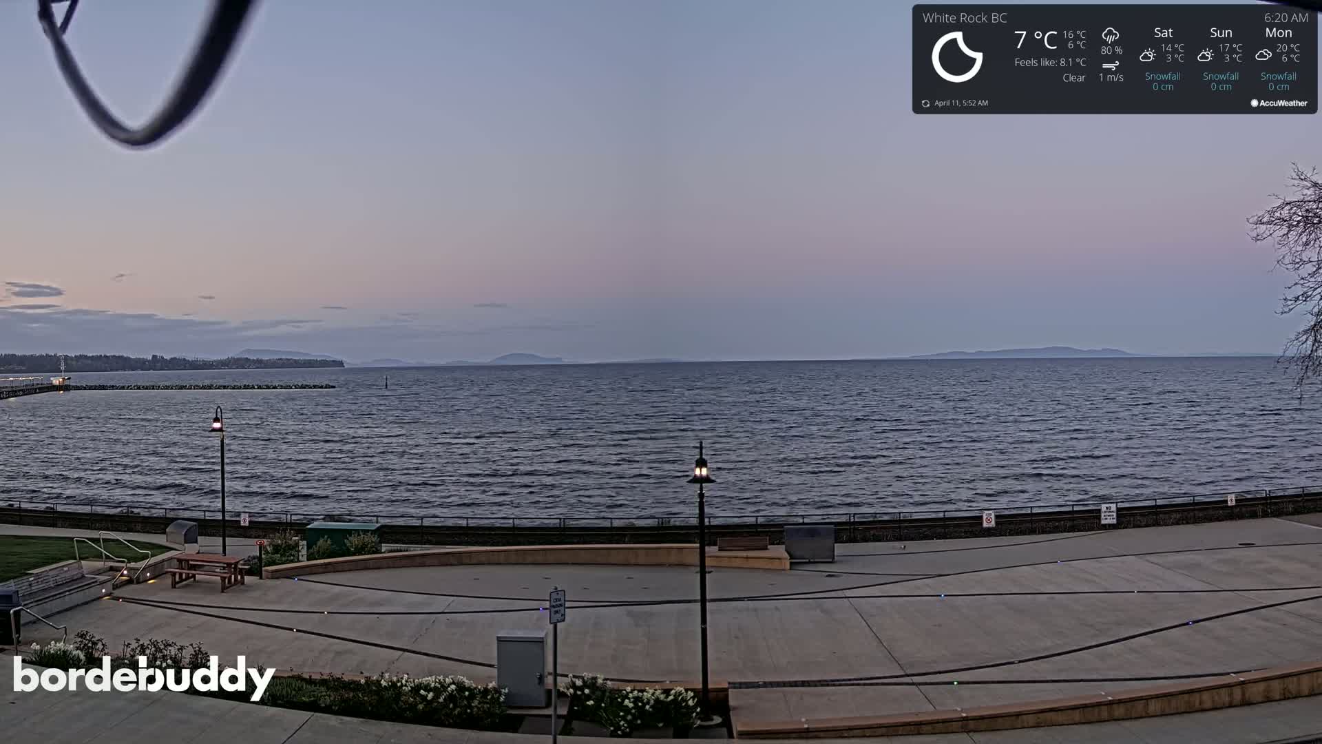 A paved waterfront area with benches and lampposts overlooks a calm, dark-blue sea under a clear, twilight sky, with distant mountains visible on the horizon.