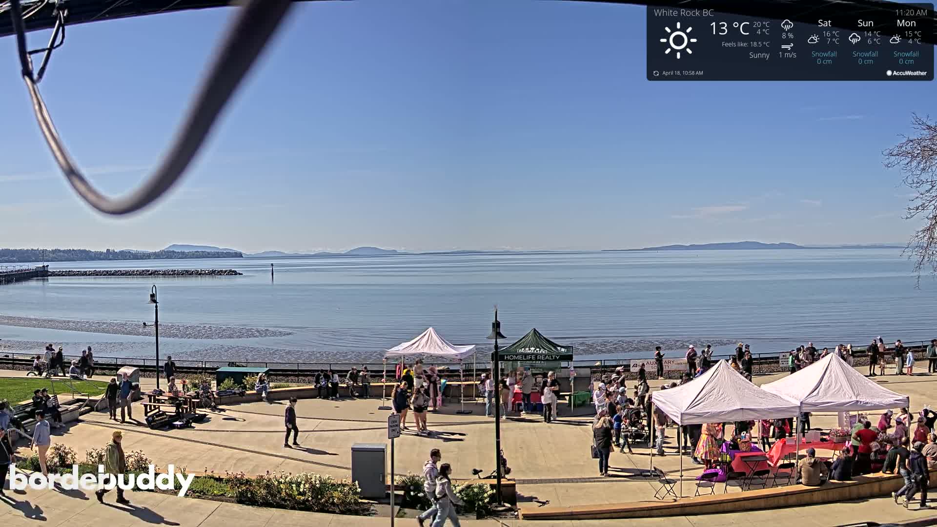 A sunny day overlooks a waterfront plaza with several market stalls and people gathered, all under a clear blue sky.