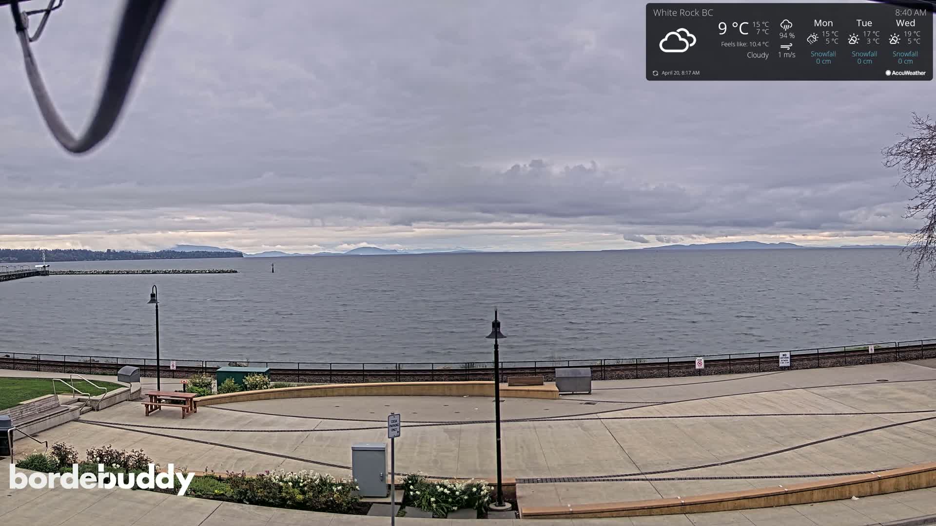 A cloudy day overlooks a calm body of water with distant mountains visible under a gray sky, and a paved waterfront area with benches and lampposts in the foreground.