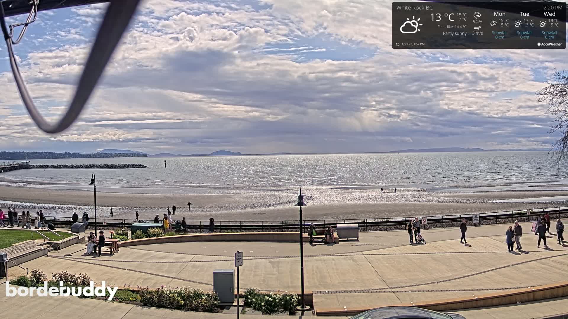 A partly sunny beach scene shows a wide sandy shore with people walking along the waterfront path and on the beach, overlooking a calm body of water and distant mountains.