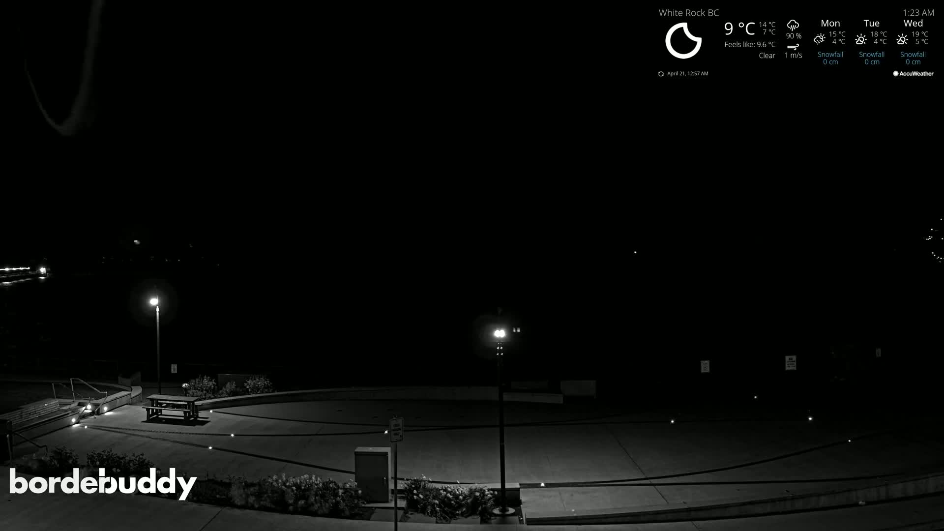 A nighttime, clear view of an empty plaza with two lit lampposts, a picnic table, and low landscaping.