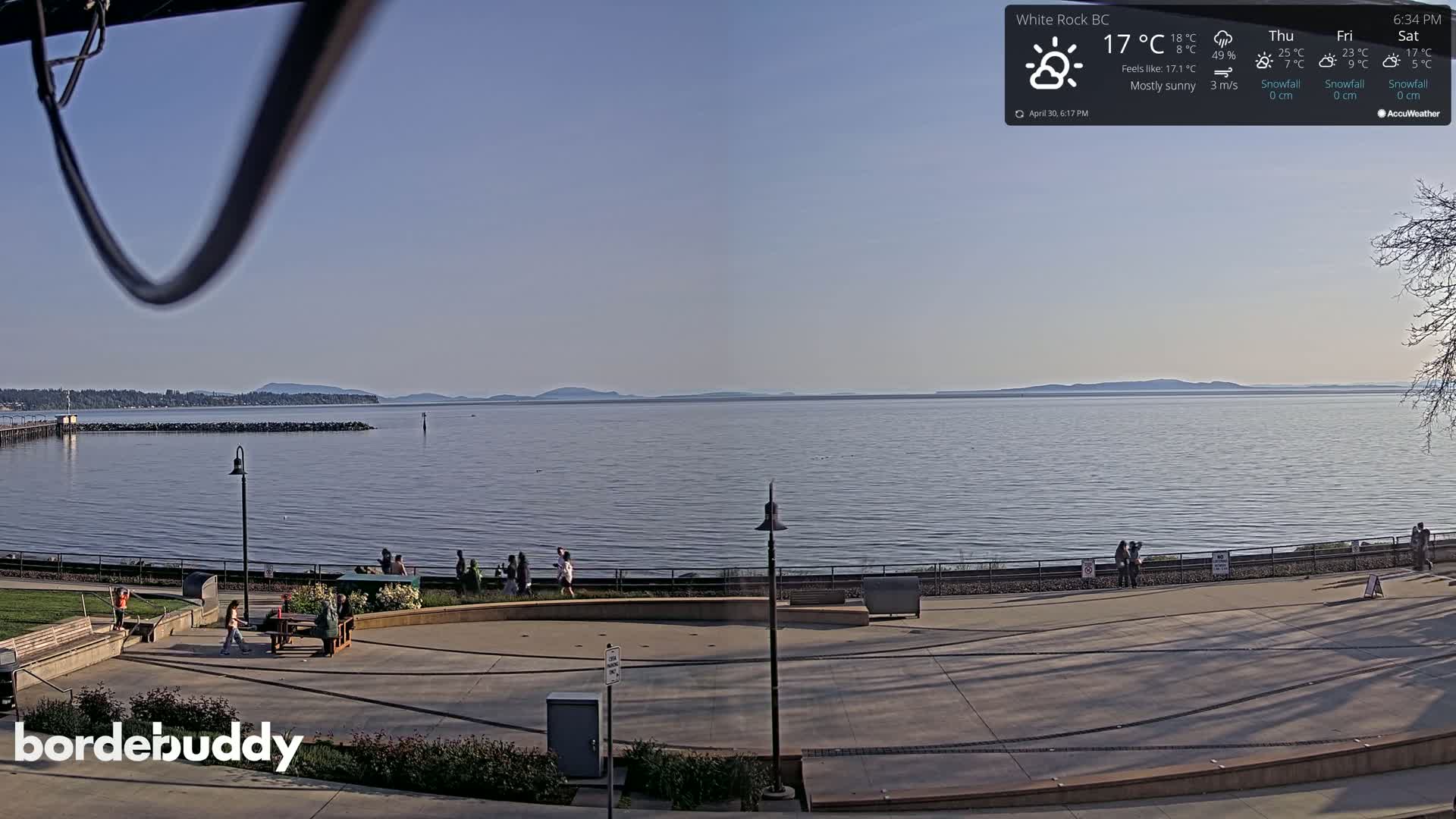 A mostly sunny waterfront scene shows a calm body of water with distant mountains under a clear sky, and people gathered in a plaza near a pier.