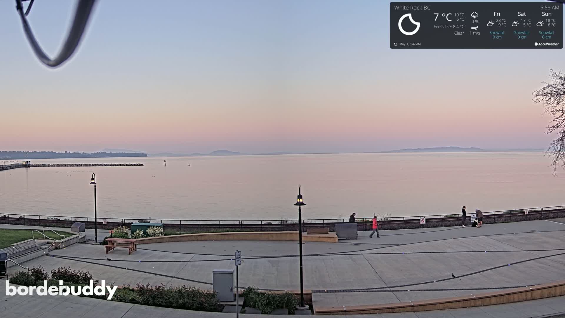 A calm, clear sunrise illuminates a paved waterfront area with a few people walking near a calm body of water and distant shoreline.