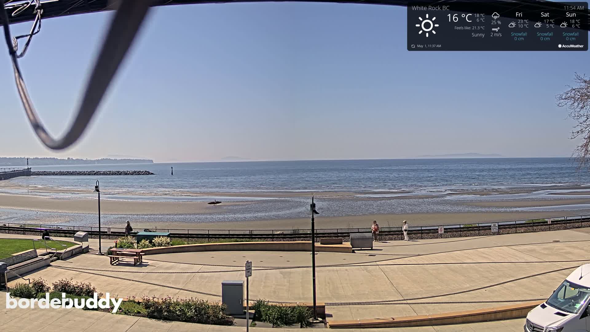 A sunny seaside paved area overlooks a calm ocean with a sandy beach at low tide, a few people walking near a fence.