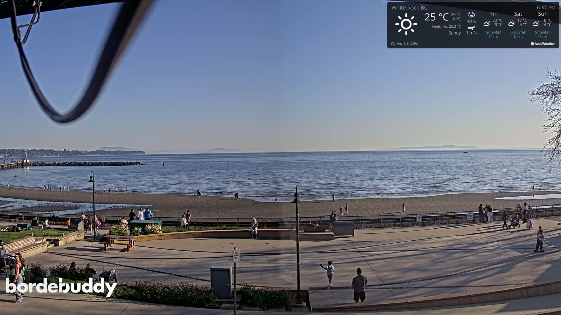 A sunny beach scene overlooks a calm ocean with distant mountains under a clear blue sky, and people are enjoying the beach and surrounding area.