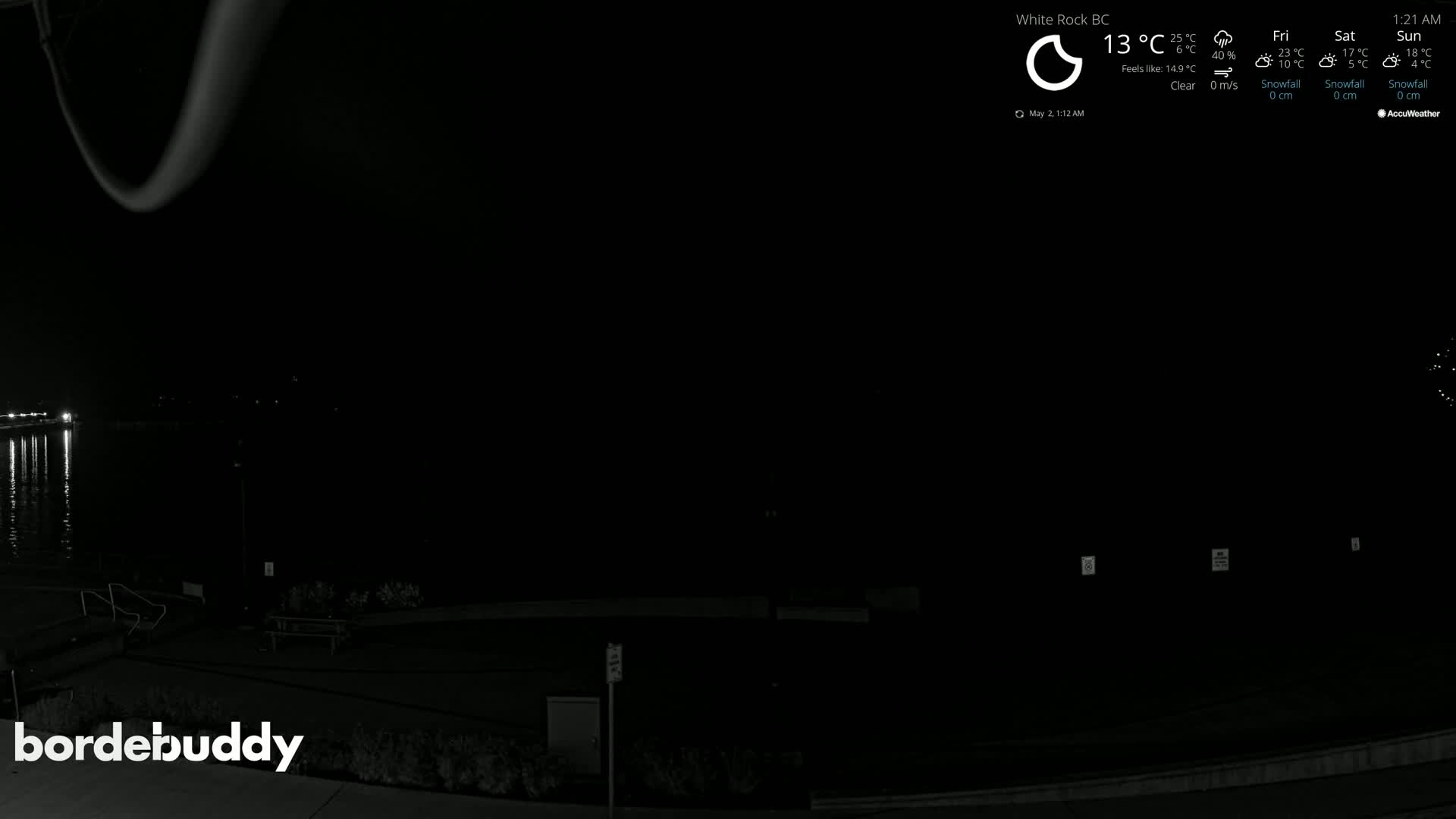 A nighttime view of a dark waterfront area with calm water reflecting lights, a paved walkway, and some park benches under a clear sky.