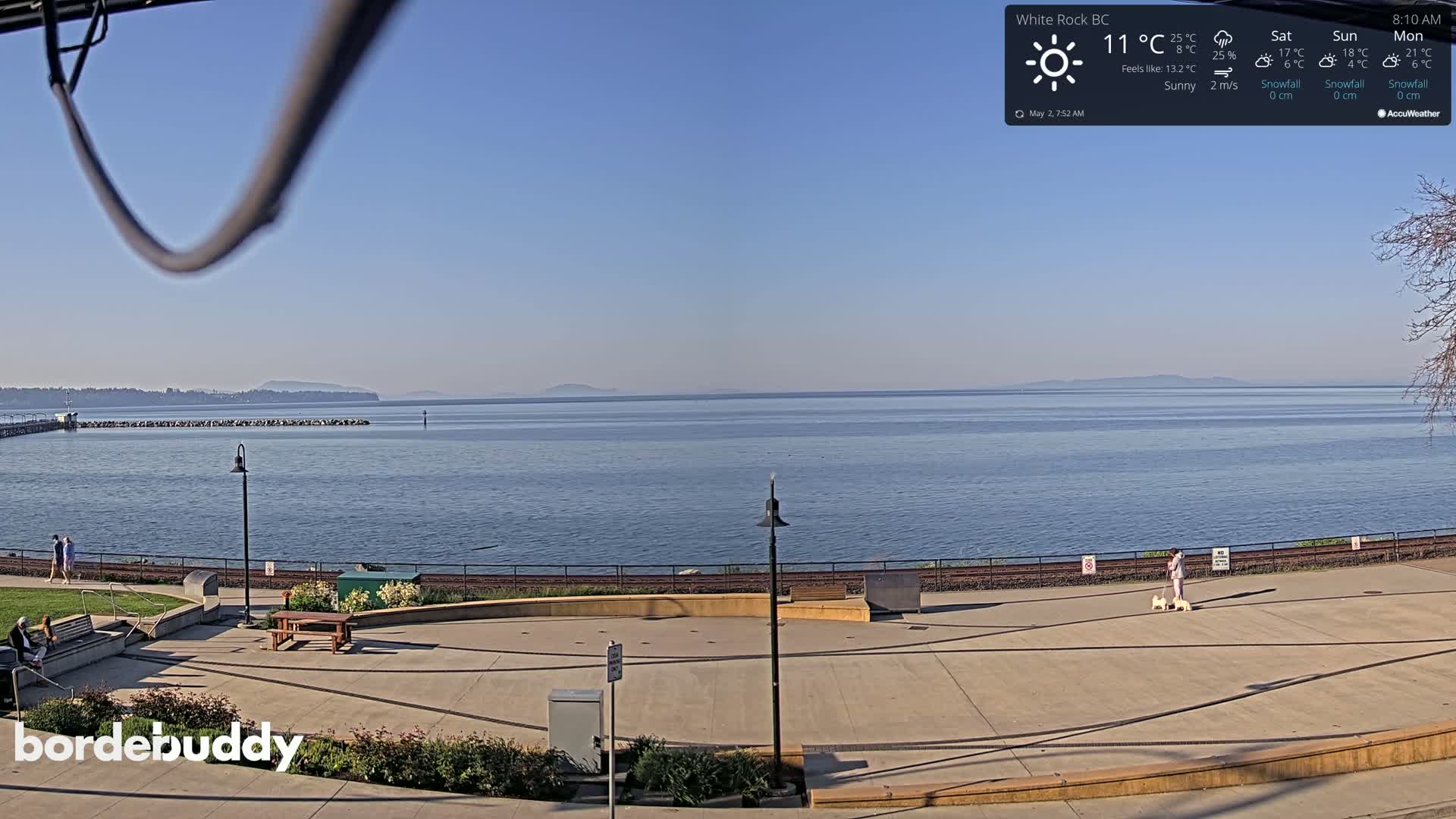 A sunny waterfront plaza overlooks a calm ocean with a distant landmass under a clear blue sky.