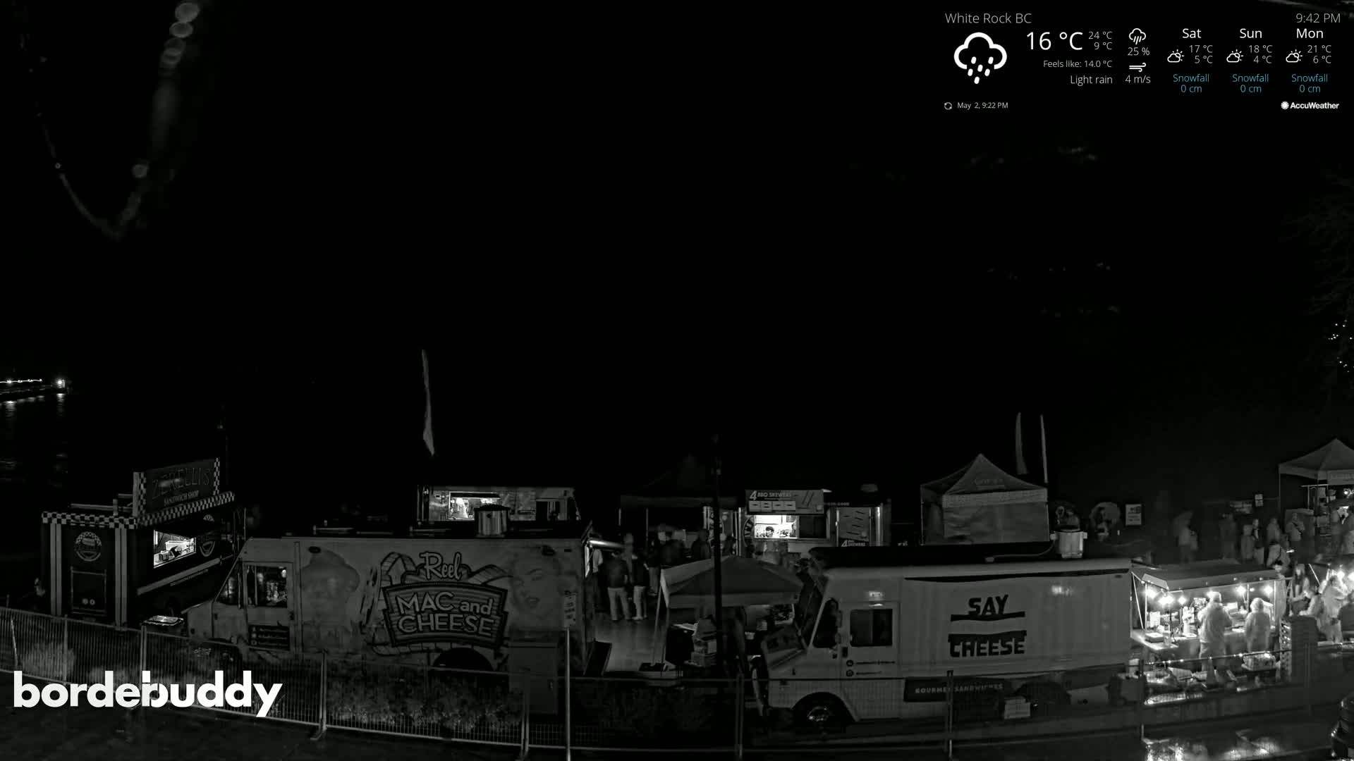 At night, under light rain, several food trucks and tents are illuminated, with people gathered around them.