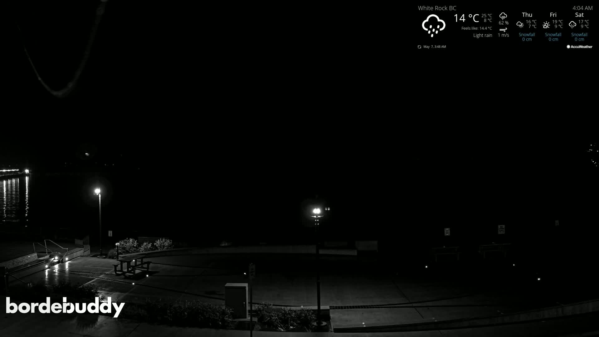 A nighttime, lightly raining scene shows an empty waterfront area with a few lights and some park benches.