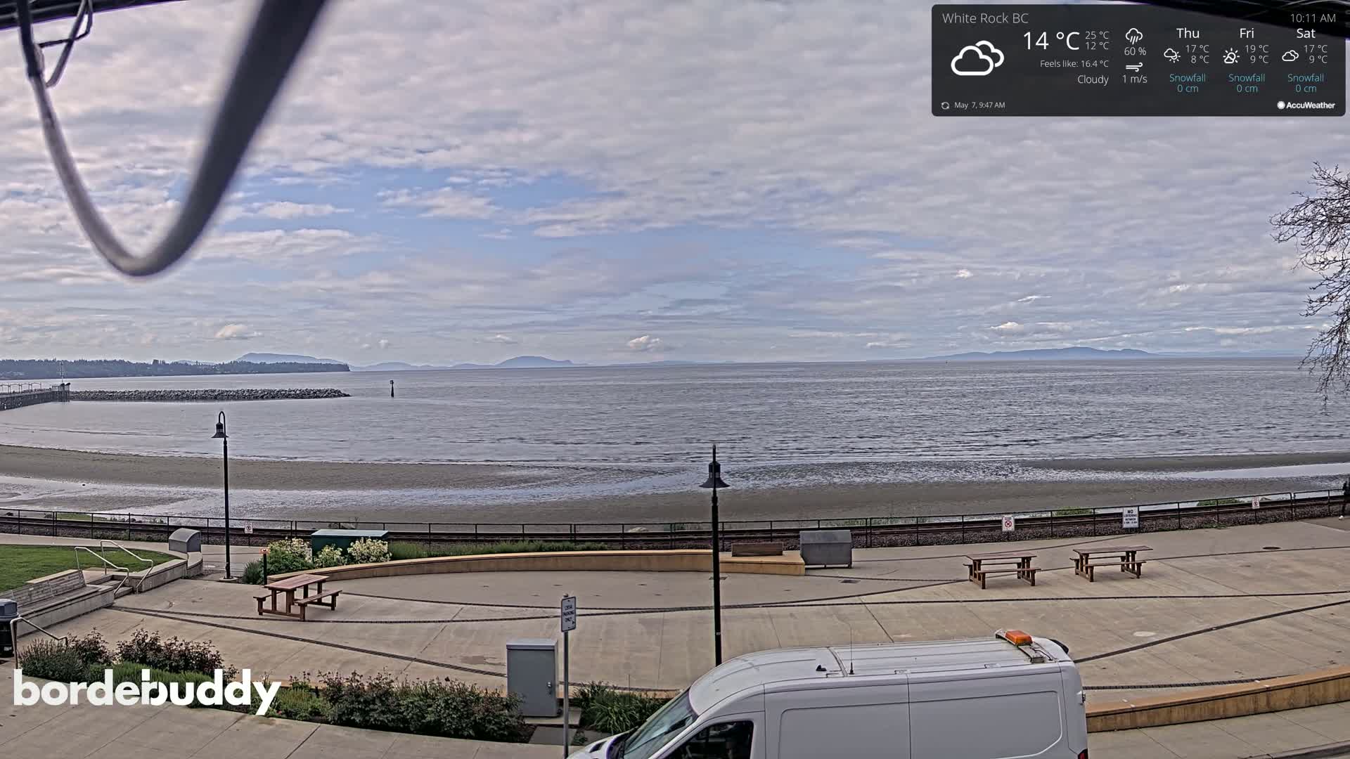 A cloudy day overlooks a calm bay with a sandy beach and paved waterfront area containing picnic tables and a van.