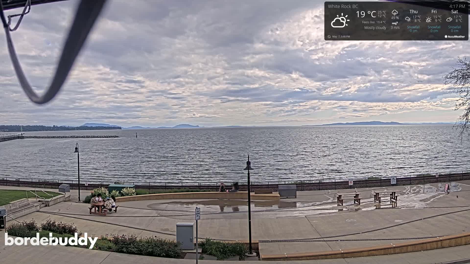A mostly cloudy day overlooks a calm body of water with distant mountains, a waterfront park with picnic tables, and several people sitting at one of the tables.