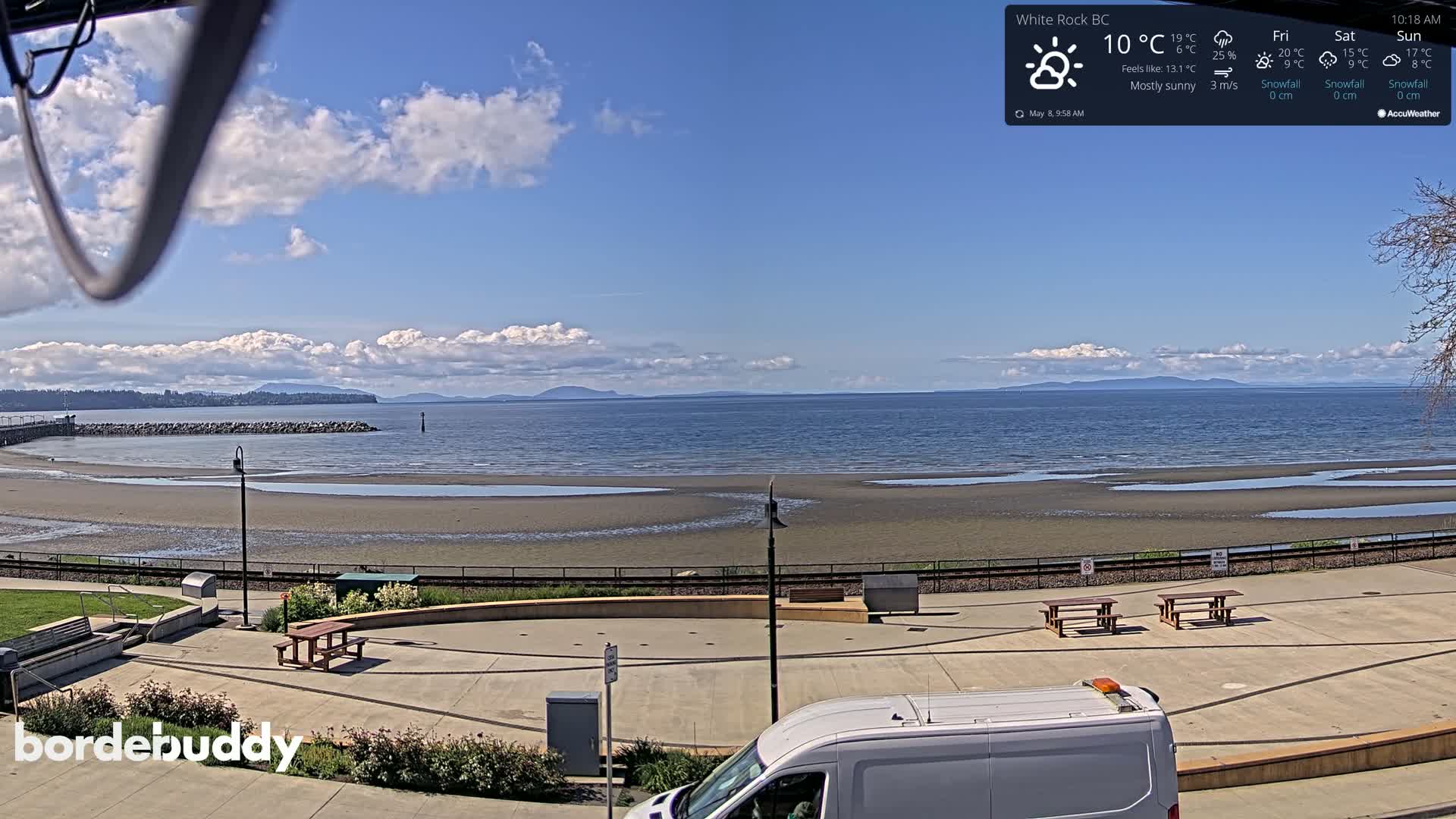 A mostly sunny day shows a view of a sandy beach with picnic tables and a van parked nearby, overlooking a calm ocean and distant mountains.
