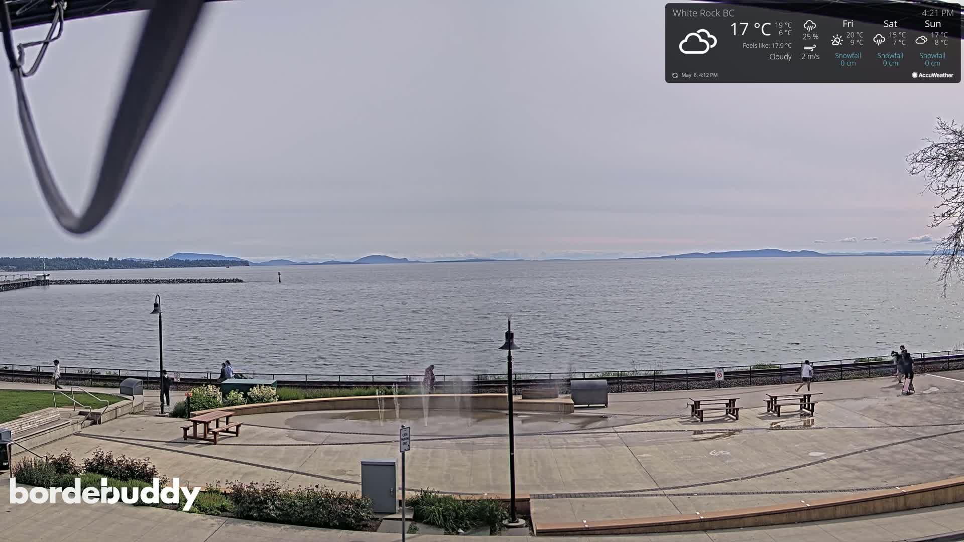 A cloudy outdoor scene shows a calm body of water with distant mountains under a mostly gray sky, and a paved waterfront area with picnic tables and small water features.