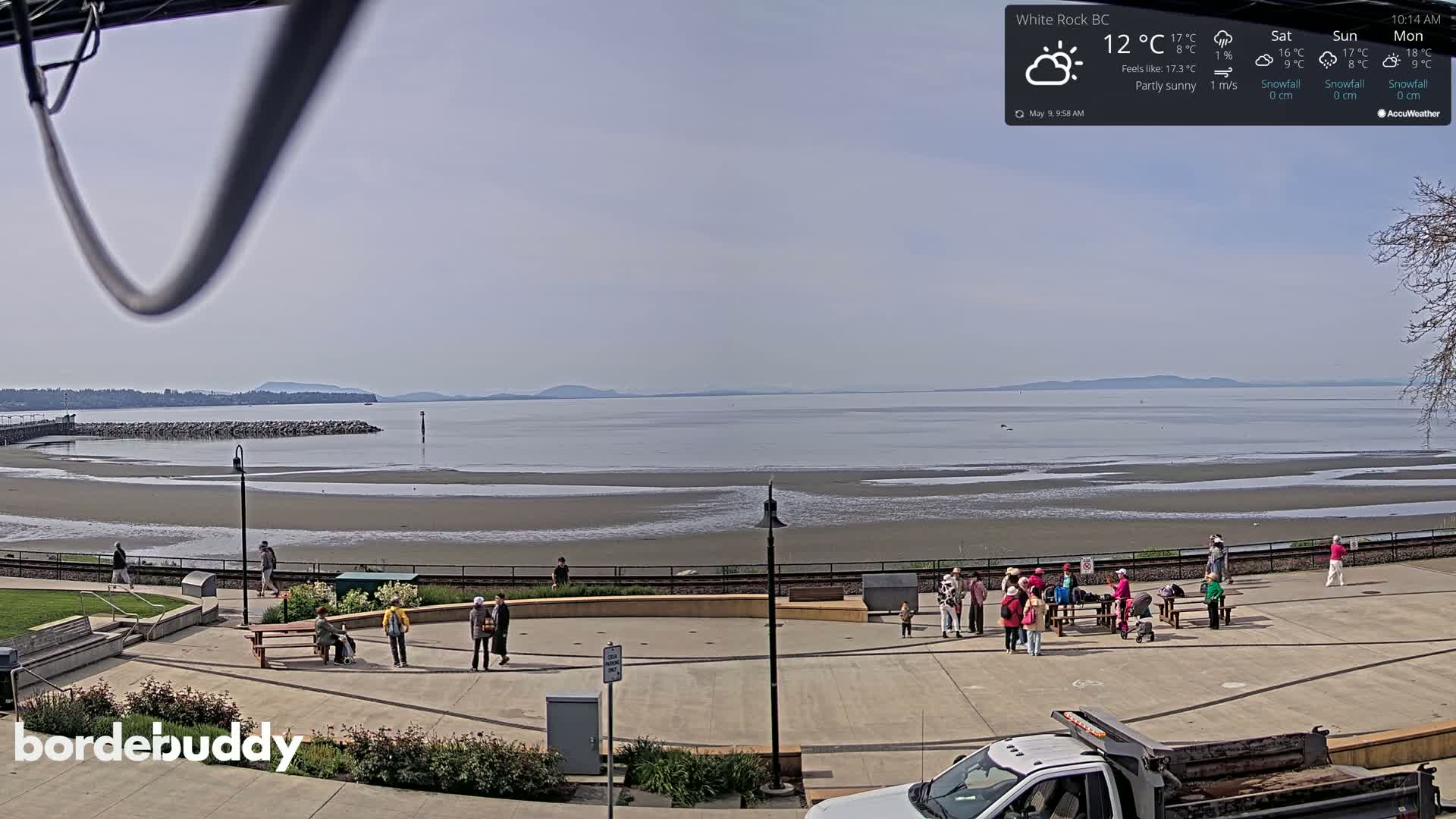 A partly sunny day shows a wide expanse of calm water and exposed mudflats at low tide, with people gathered in a paved plaza near a waterfront park.
