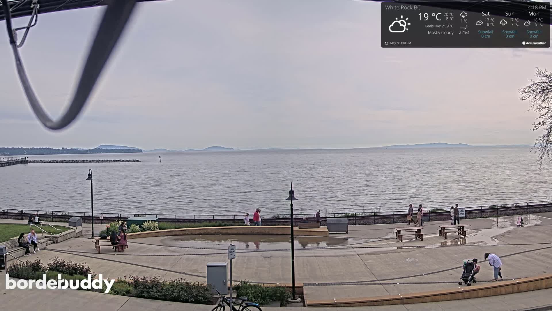 A mostly cloudy day shows a calm body of water with distant mountains, a waterfront park with people, and wet pavement.