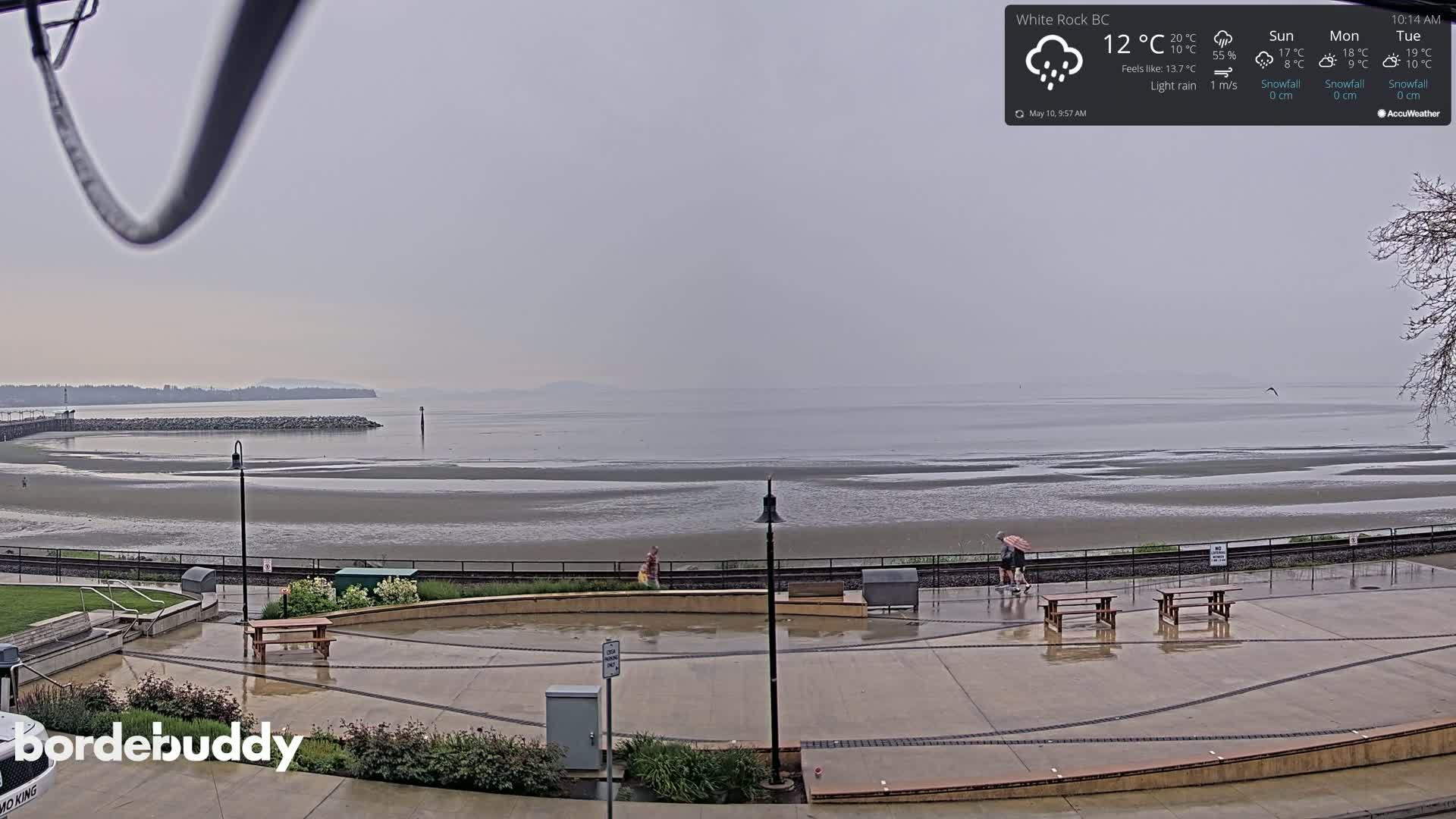 A wet, paved plaza overlooks a wide, exposed mudflat and calm body of water under a gray, overcast sky.