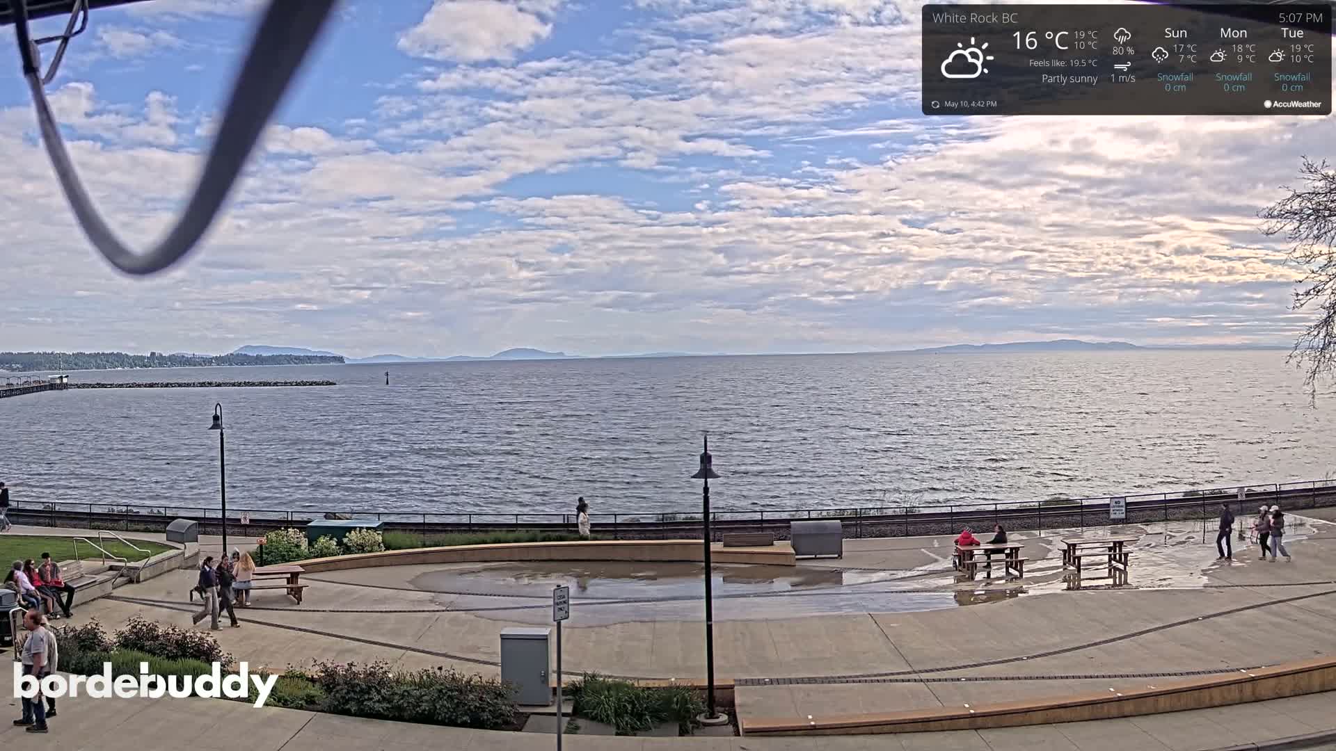 A partly sunny waterfront scene shows a paved plaza with several people and picnic tables near calm water, beyond which is a distant mountain range under a mostly cloudy sky.