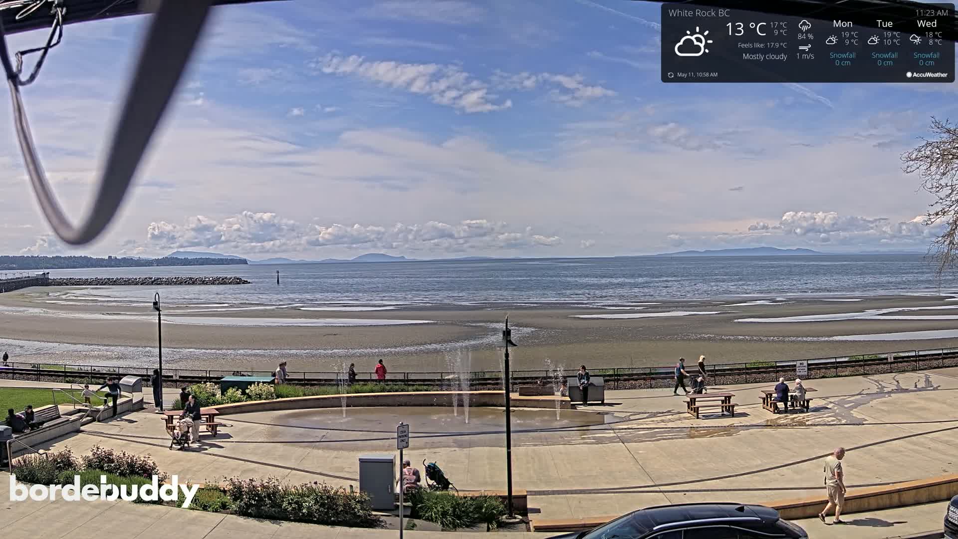 A mostly cloudy day reveals a beach with people relaxing near a water fountain, all overlooking a calm ocean and distant mountains.