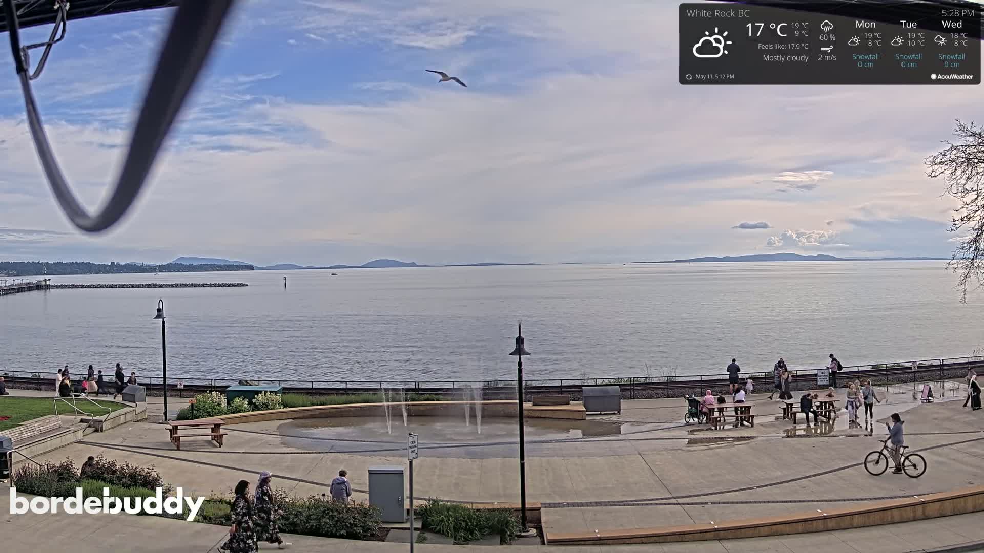 A mostly cloudy day overlooks a calm body of water with people gathered on a paved waterfront area featuring a small water fountain.