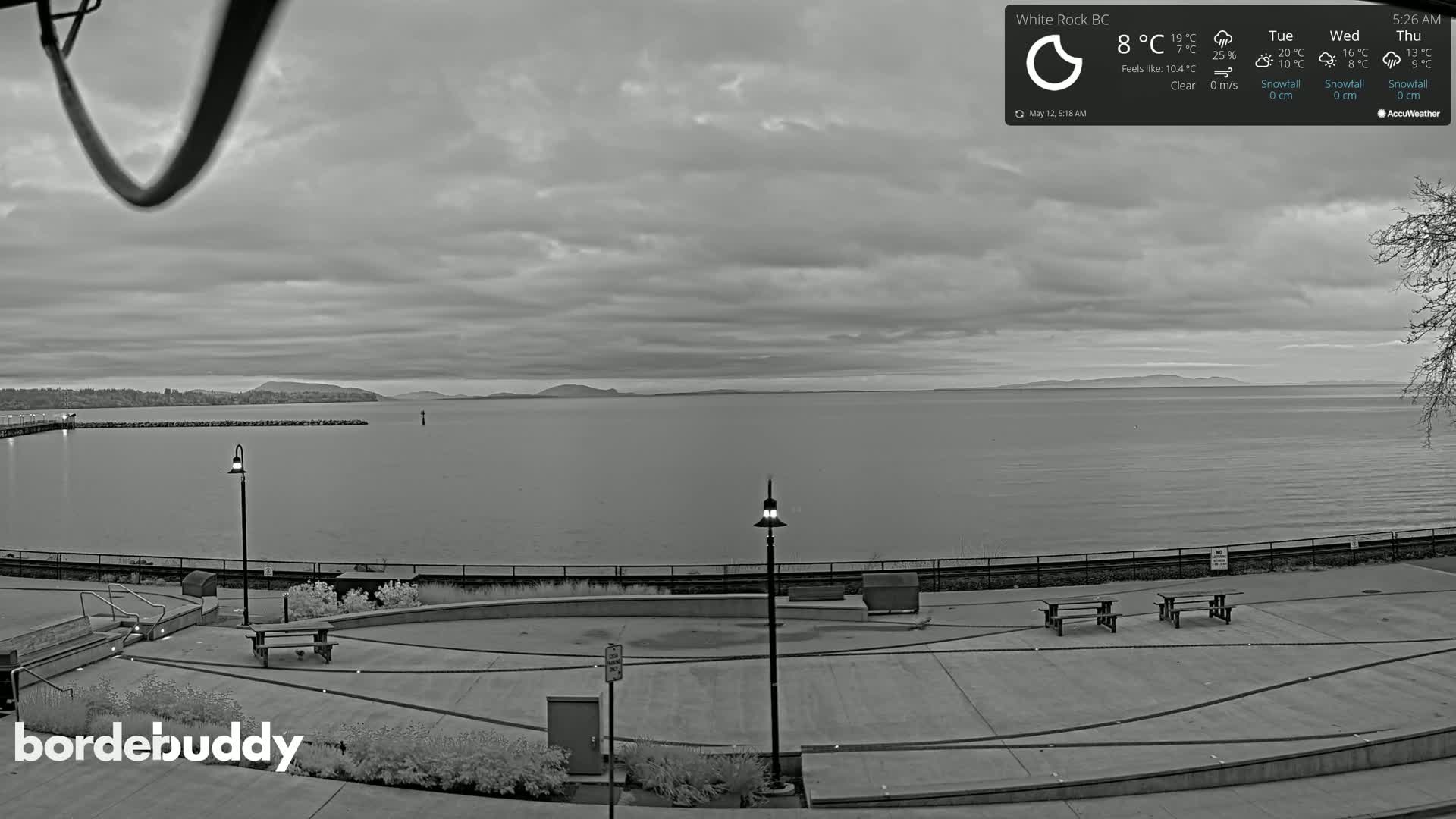 A grayscale view of a calm body of water under a cloudy sky, overlooking a paved waterfront area with benches and lampposts.