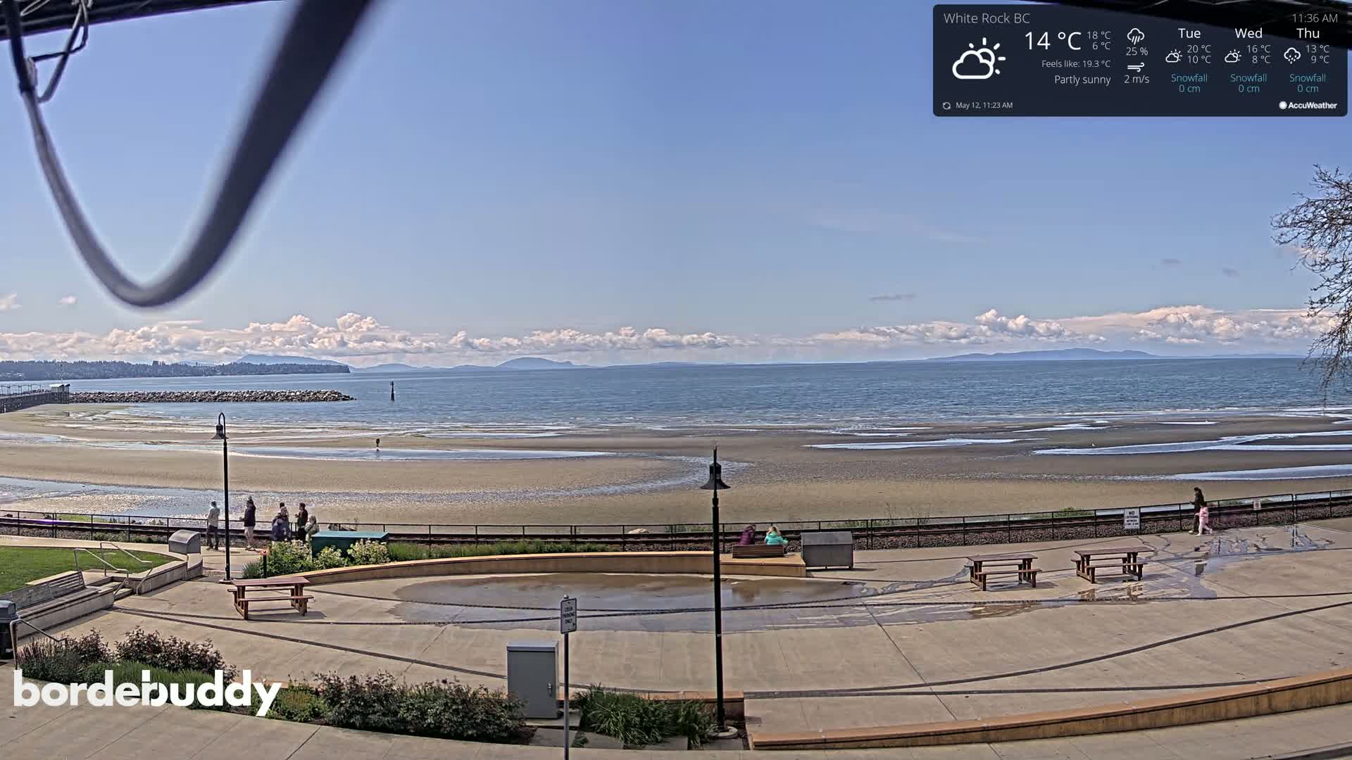 A partly sunny beach scene with a calm ocean, distant mountains, and a few people scattered on the sandy shore and paved area.