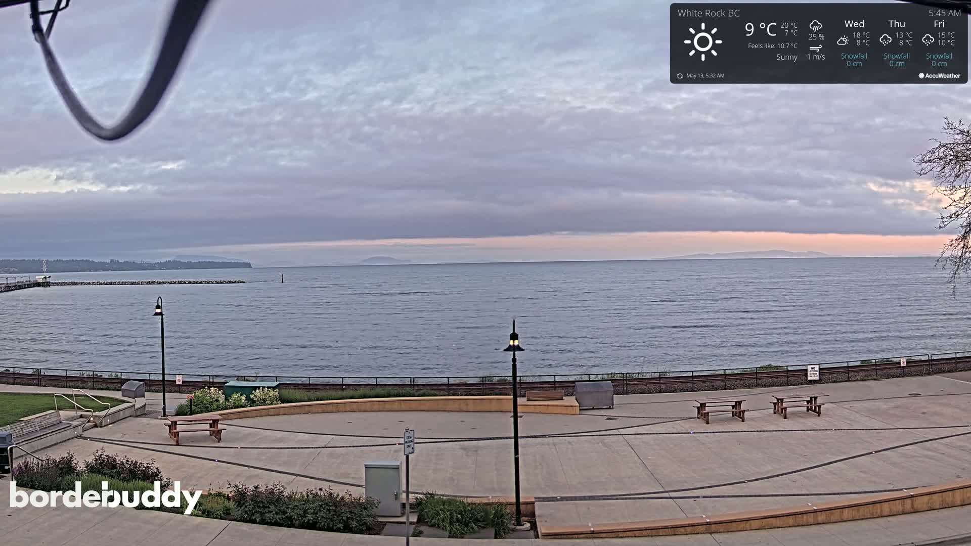 A mostly sunny, calm day overlooks a seaside plaza with benches and lampposts, a calm ocean, and distant land.