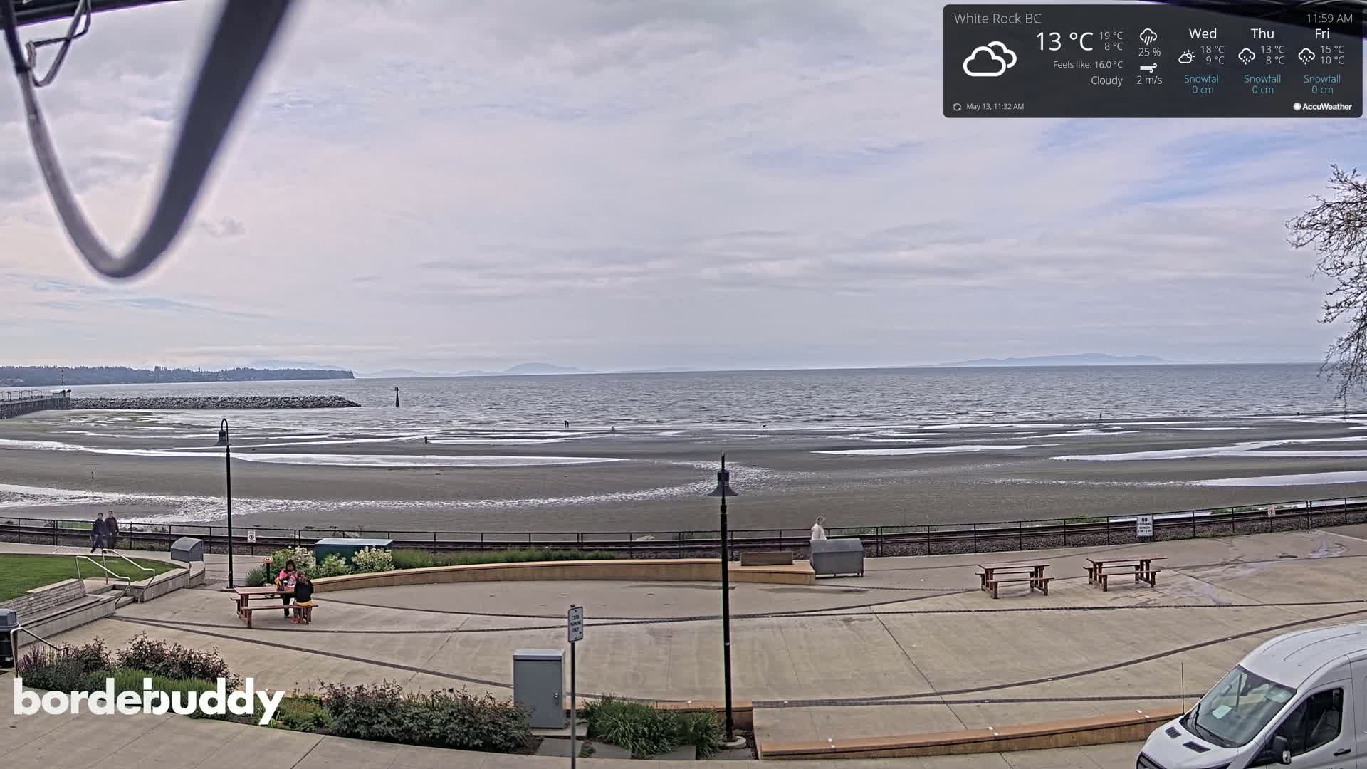 A cloudy day at a beach with a wide expanse of calm water and a paved walkway with picnic tables.