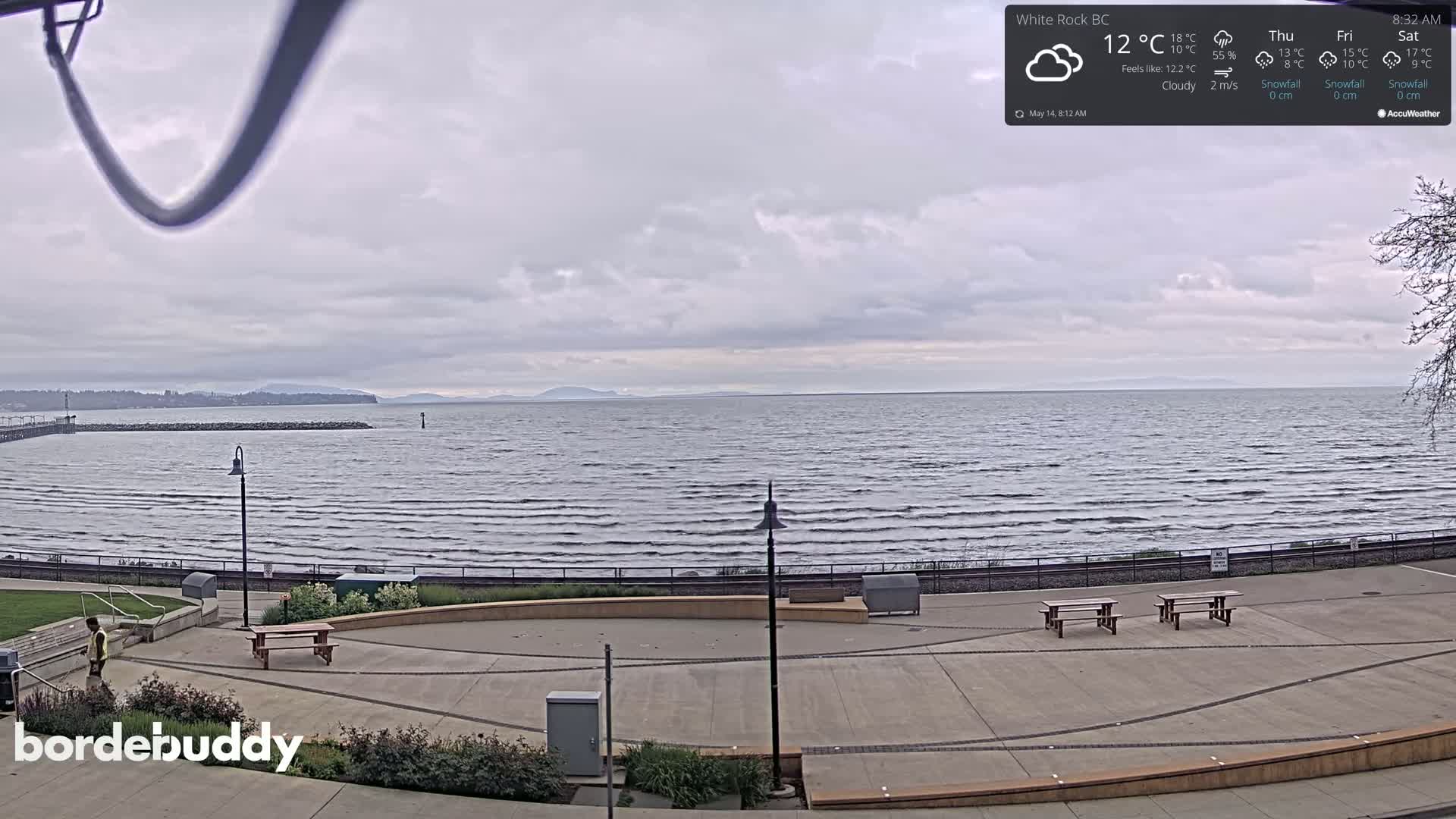 A cloudy day overlooks a calm body of water with a paved waterfront area containing picnic tables and a person walking.