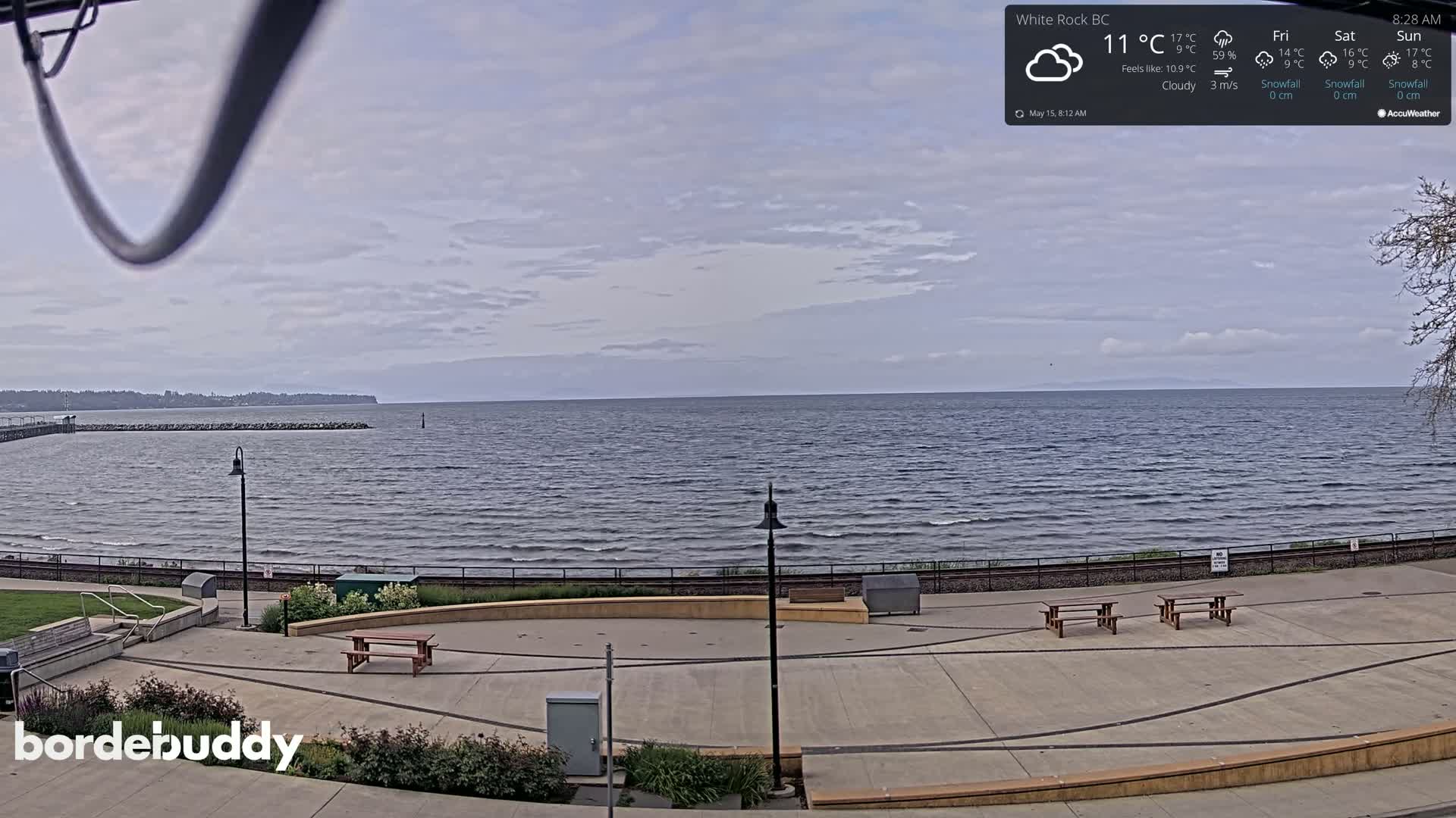 A cloudy day shows a calm sea with a paved waterfront area containing picnic tables and lampposts.