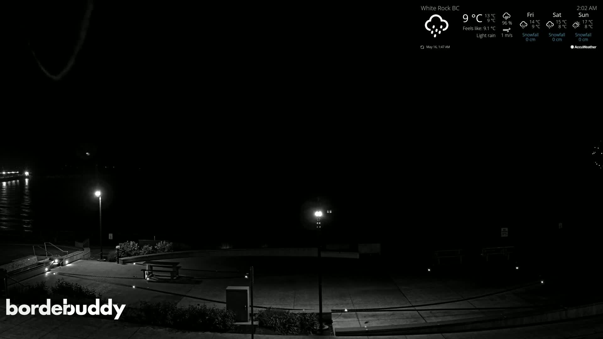 A nighttime, lightly raining scene shows an empty plaza with benches and lampposts near a body of water.