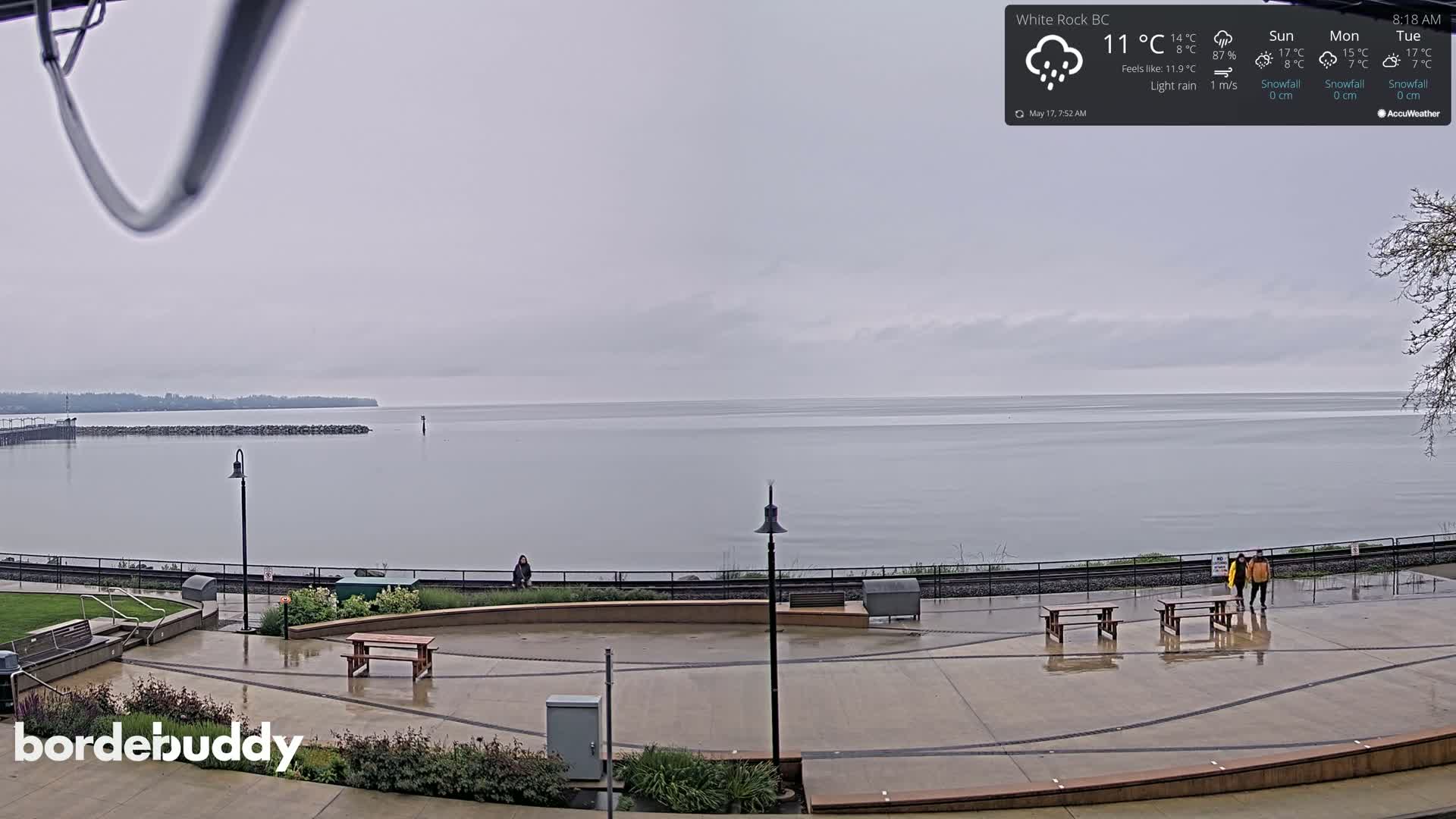 A wet, paved waterfront area with picnic tables and a few people, overlooks a calm, gray sea under an overcast sky.