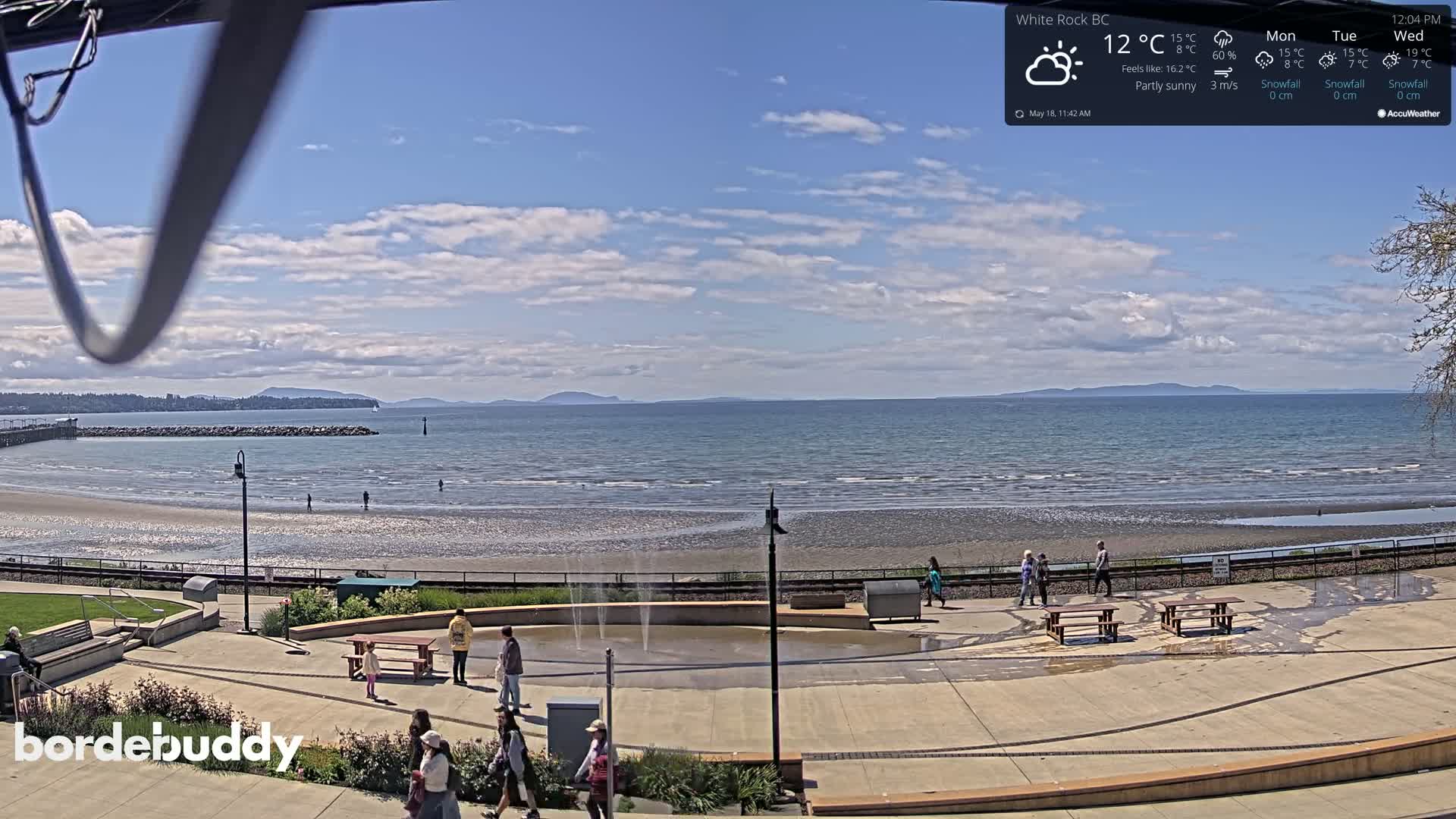 A partly sunny day shows a beach scene with people walking near a water fountain and picnic tables, overlooking a calm ocean and distant mountains.