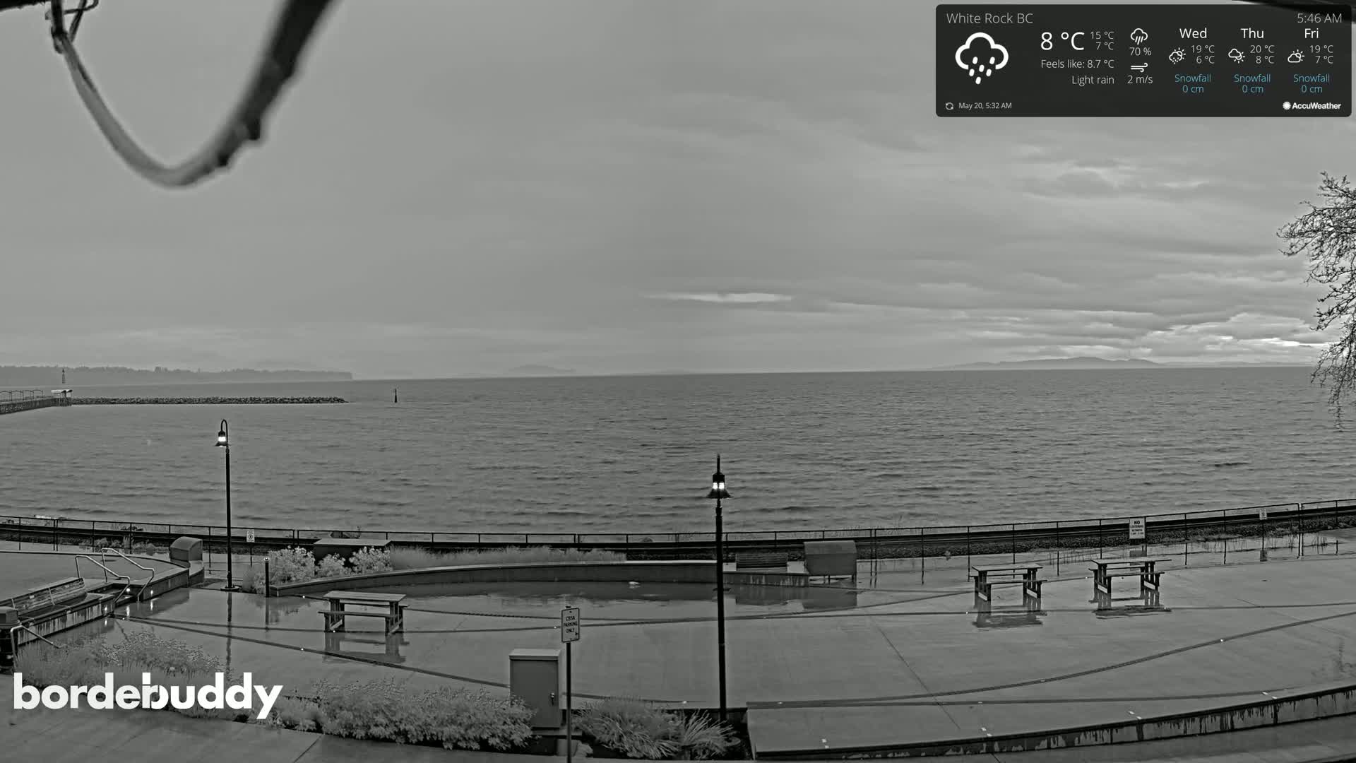 A wet, gray day is shown overlooking a calm body of water with a distant shoreline and a paved waterfront area with picnic tables.