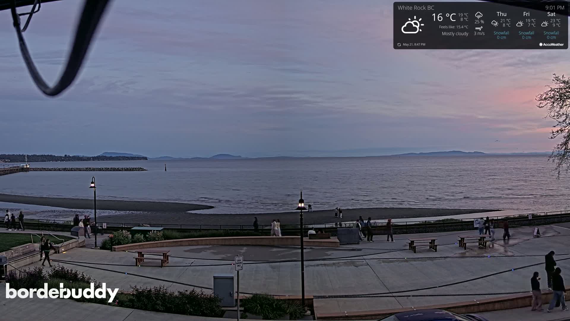 A mostly cloudy twilight scene shows a calm body of water with a beach and a paved plaza with people, picnic tables, and lights, viewed from an elevated perspective.
