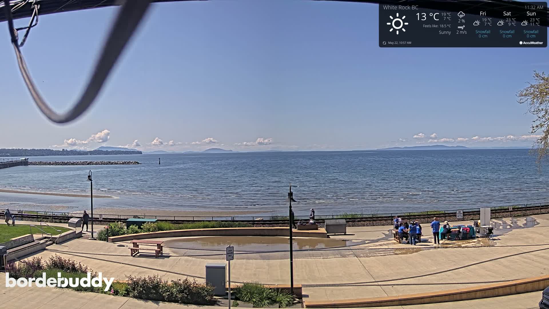 A sunny seaside view shows a calm ocean with distant mountains under a mostly clear sky, and a paved area with people gathered near picnic tables.