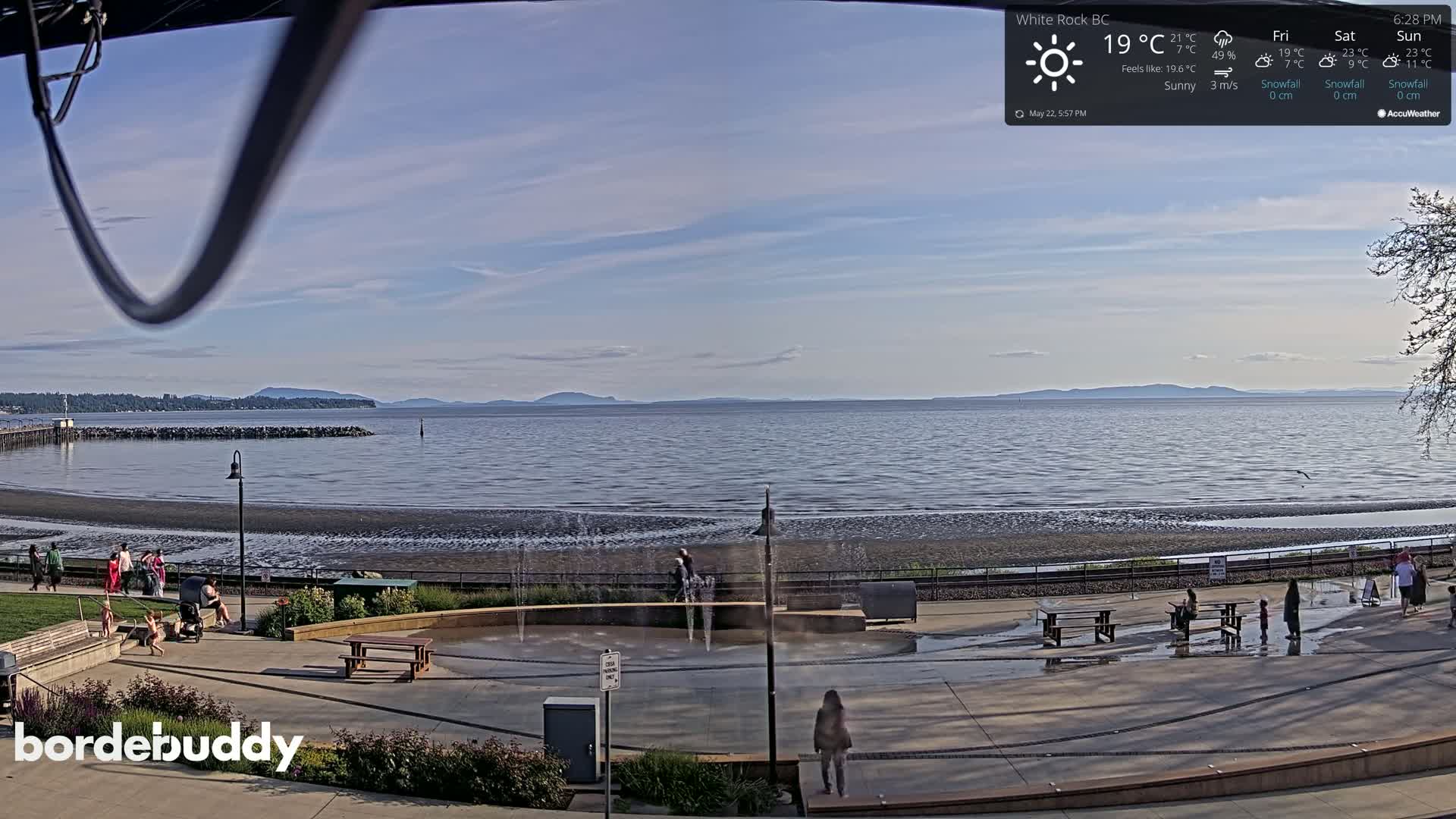 A sunny day at a waterfront park shows a calm body of water with distant mountains under a mostly clear sky; people are visible near a water feature and walking along a paved area.