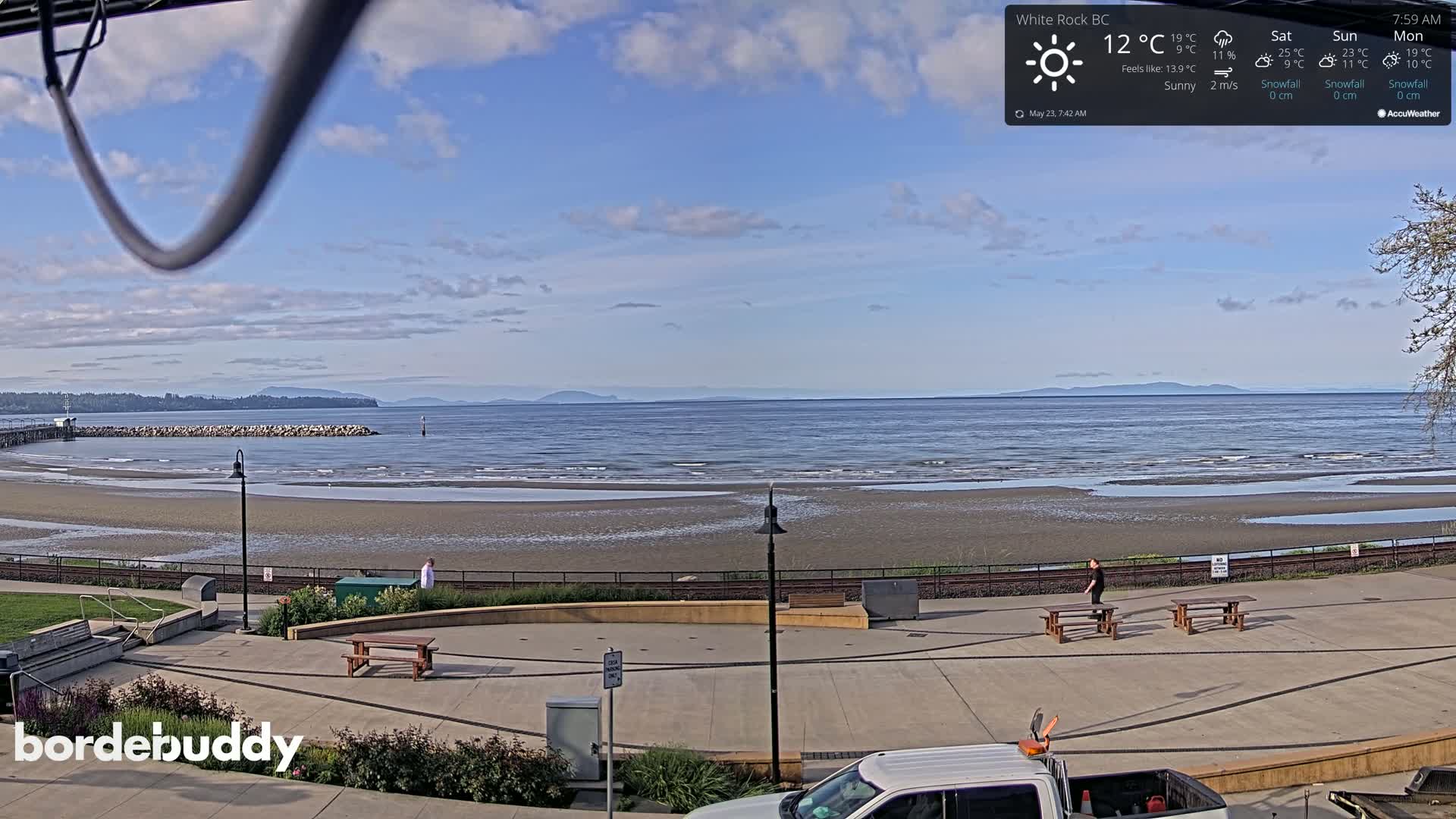 A sunny beach with calm ocean waves, a paved walkway with picnic tables, and distant mountains under a partly cloudy sky.